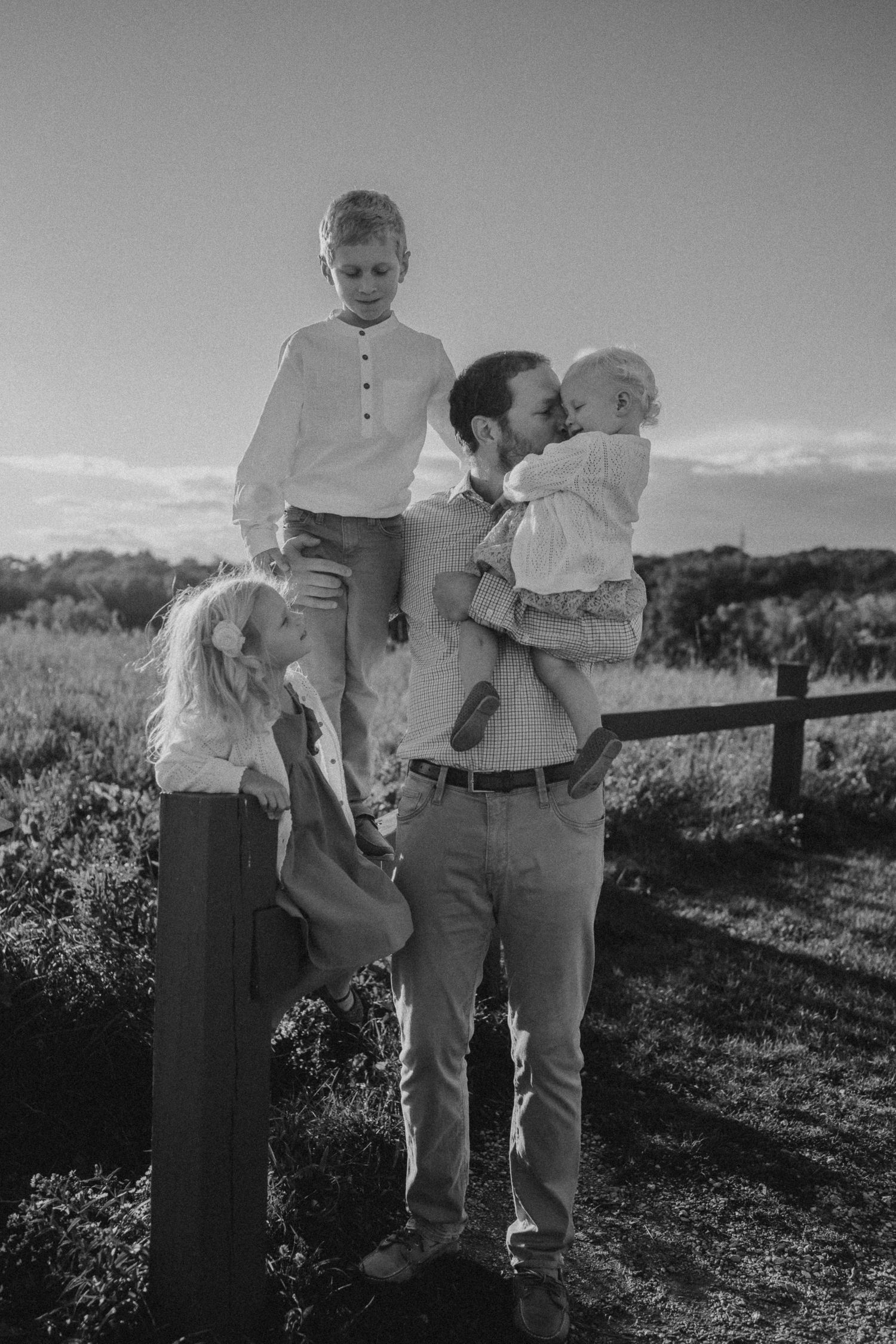 Black and white family photo taken at sunset with a wooden fence and scenic landscape in the background.