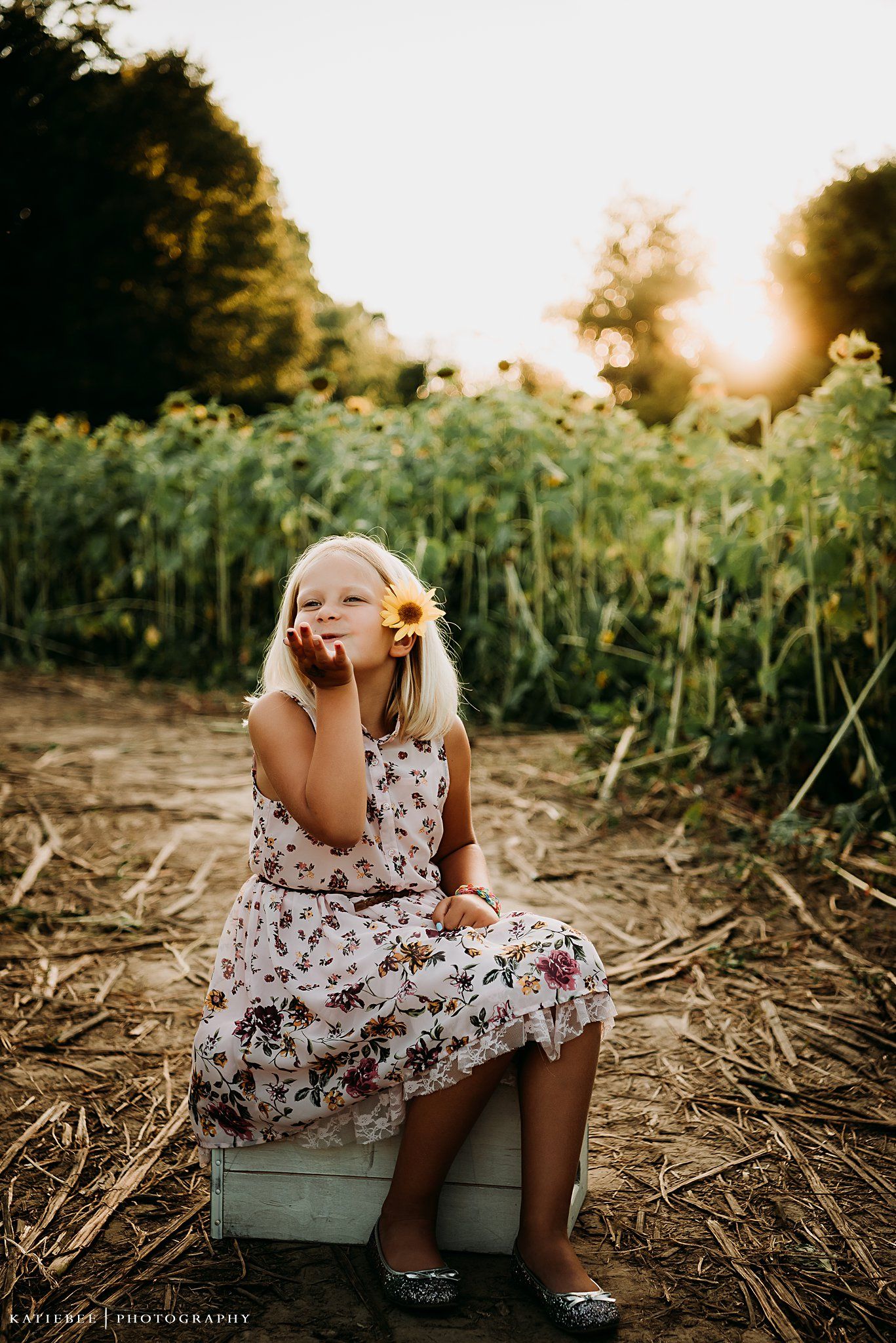 Young child in floral dress sitting thoughtfully on wooden crate among sunflowers at sunset.