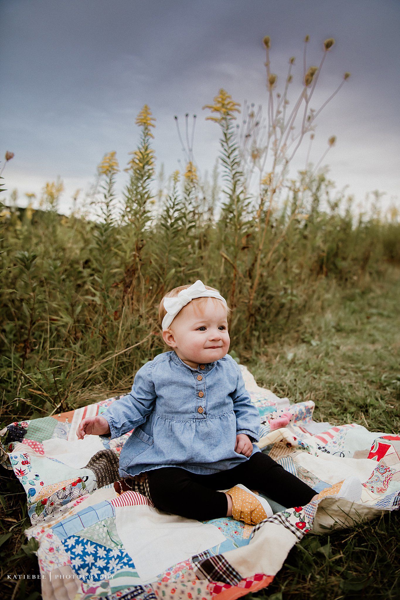 Baby in denim dress sits on colorful patchwork quilt surrounded by wildflowers in meadow.