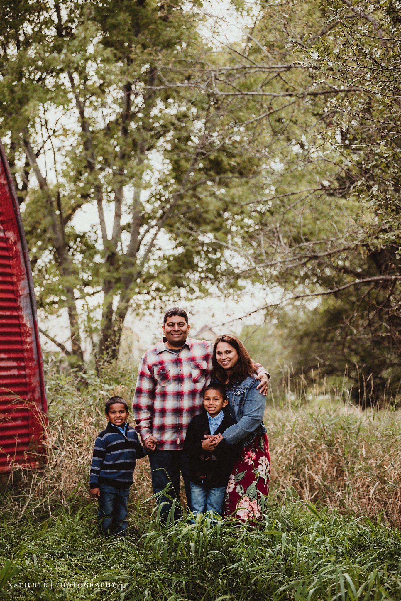 Family portrait in front of red barn with tall grass and trees in background.