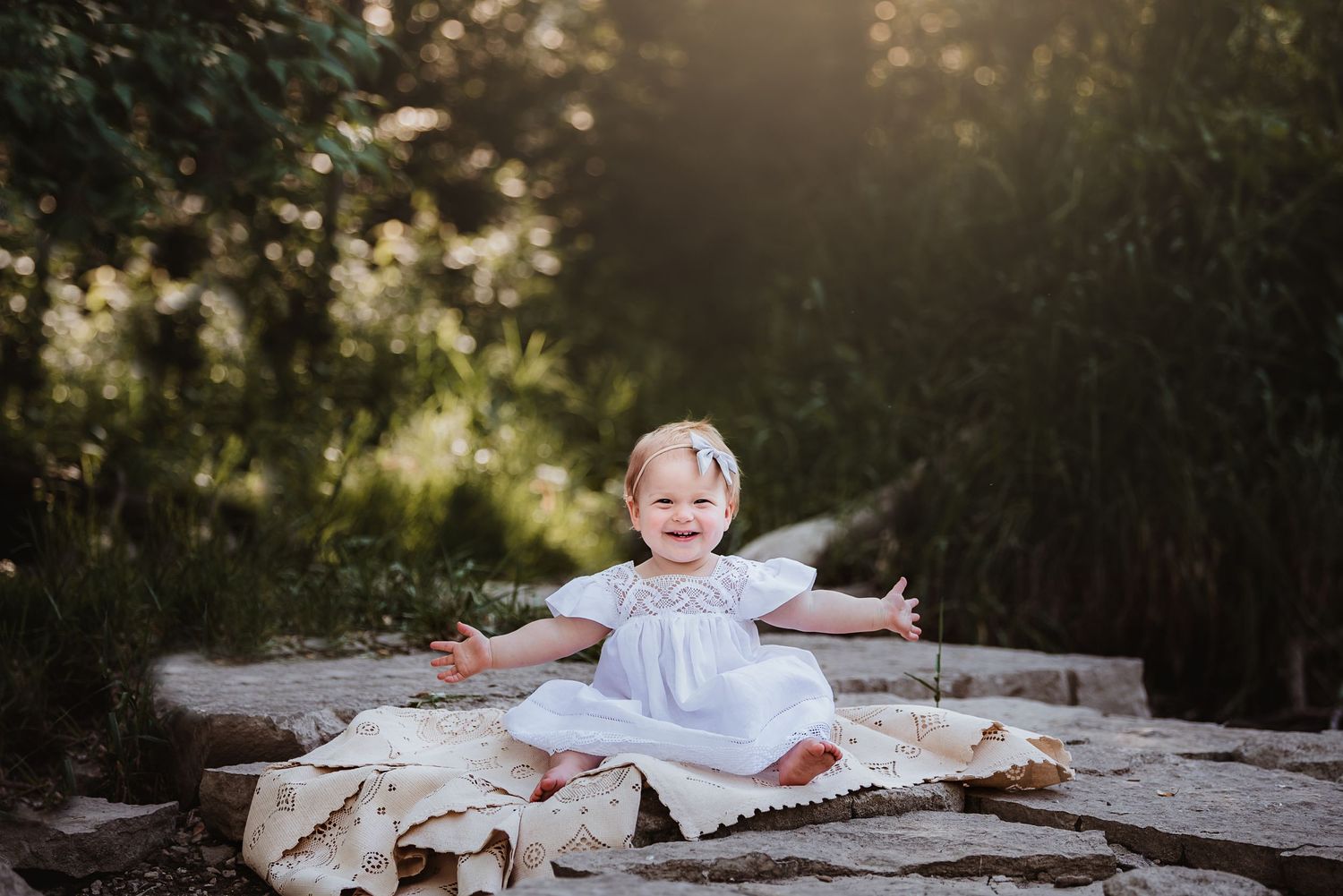 Baby in white dress sitting on white lace blanket outdoors with joyful expression.