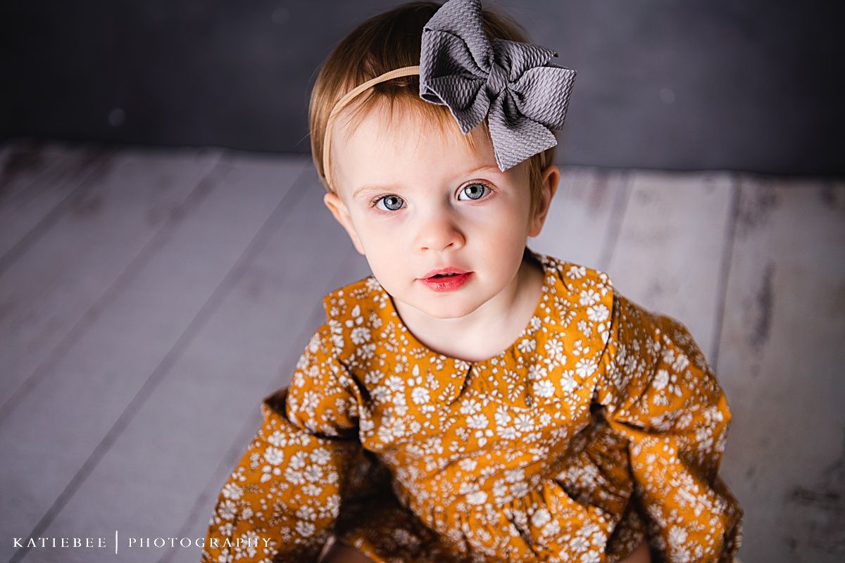 A close-up portrait showing a mustard floral dress and grey bow against dark background.