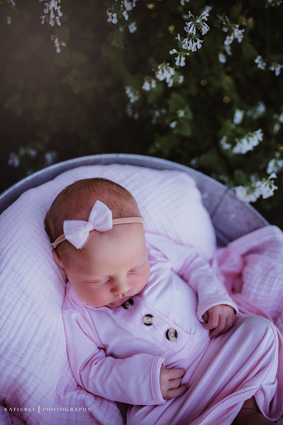 Sleeping baby in pink outfit with bow headband resting in white cushioned basket.