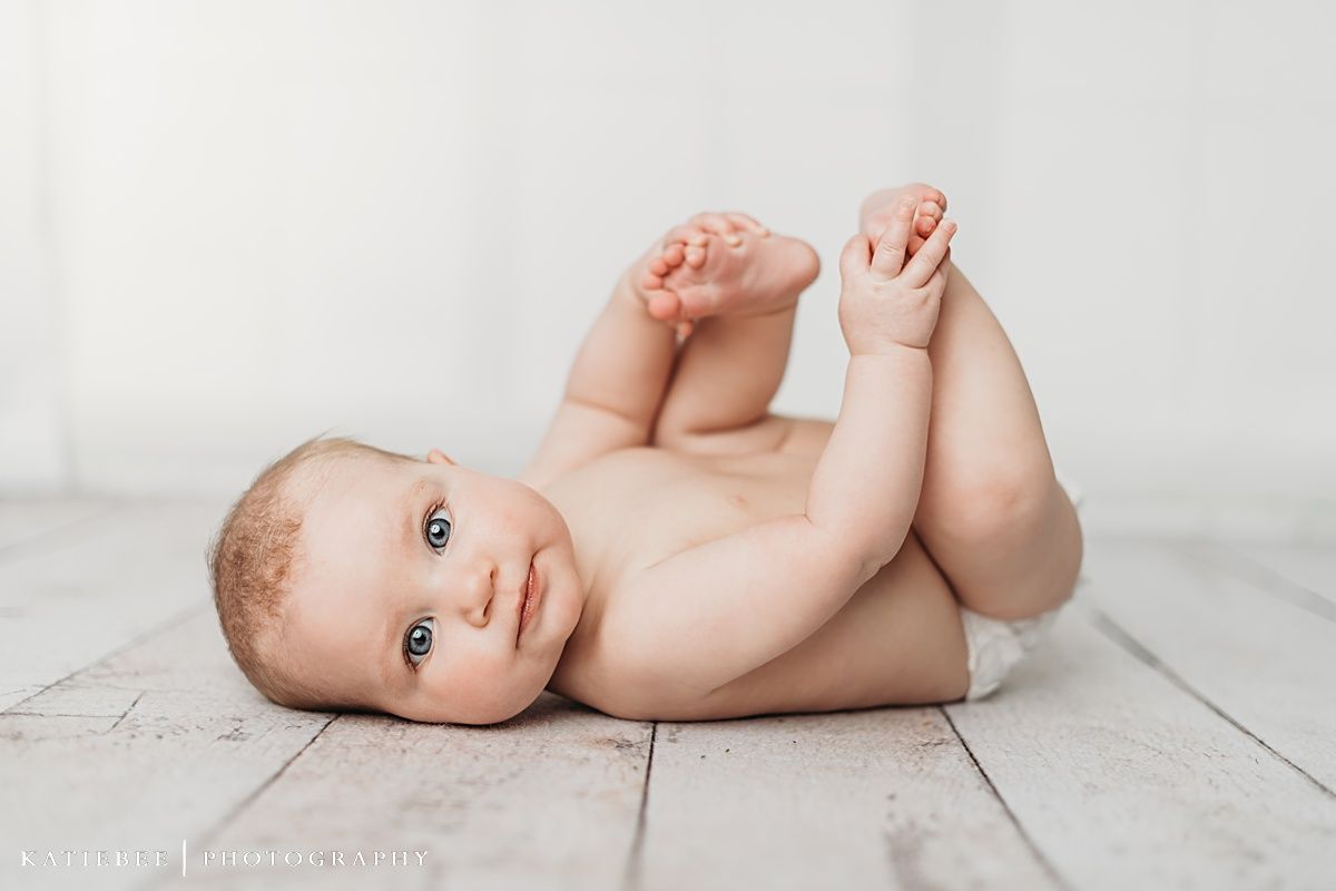 Baby lying on back on white wooden floor holding feet during photo session.