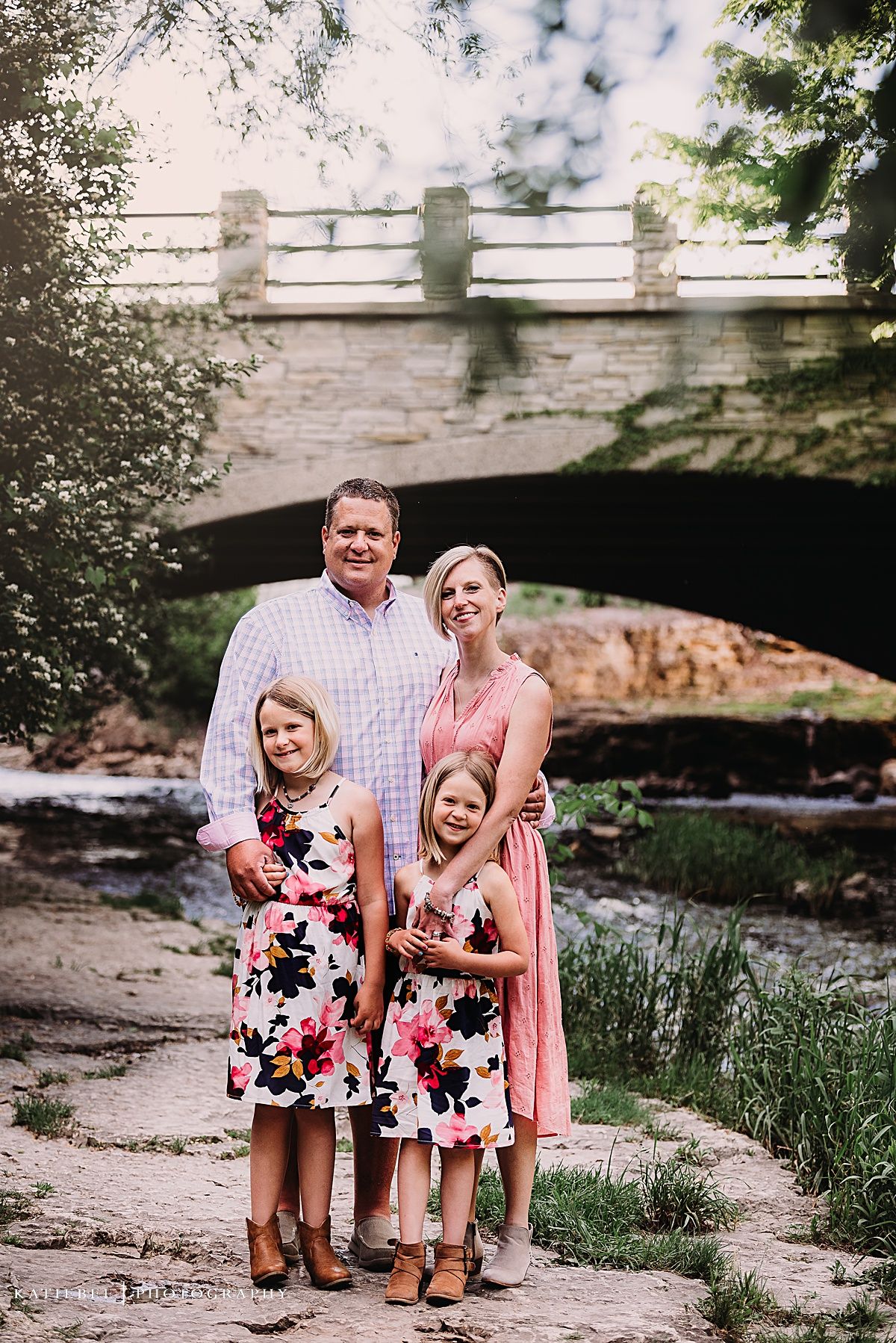 Four people dressed in coordinating outfits stand beneath a stone bridge by a creek.