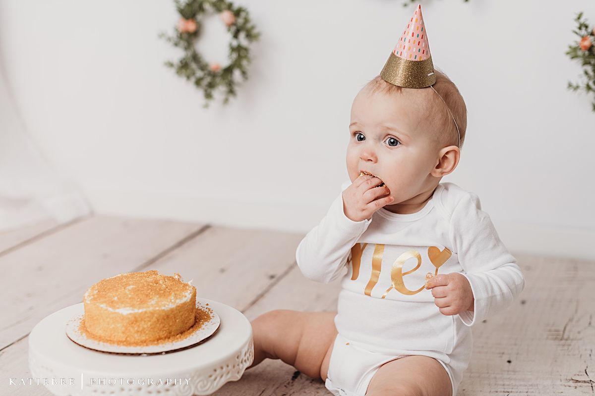 Baby in party hat reaching for cake on white pedestal with floral wreaths in background.