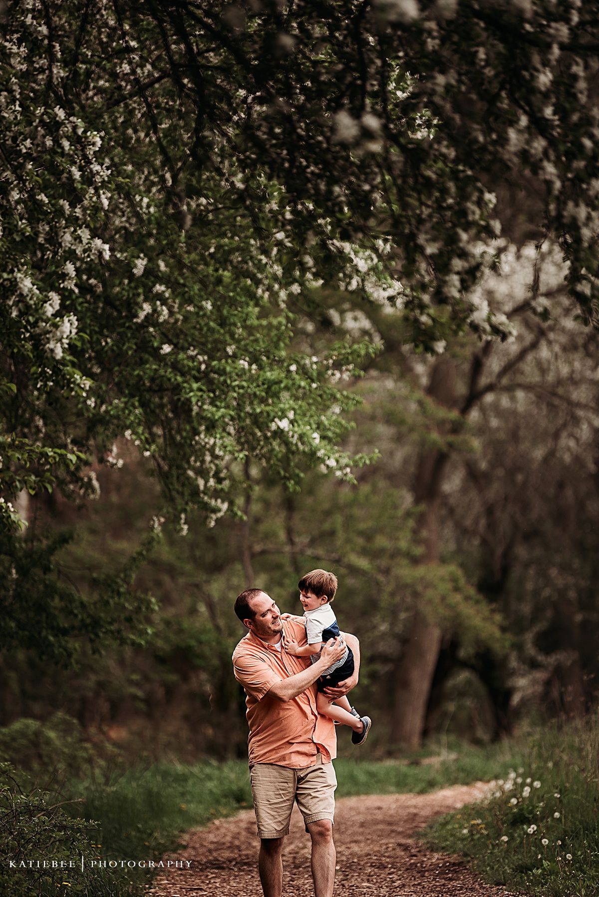A tender moment captured on a wooded path with flowering trees overhead.