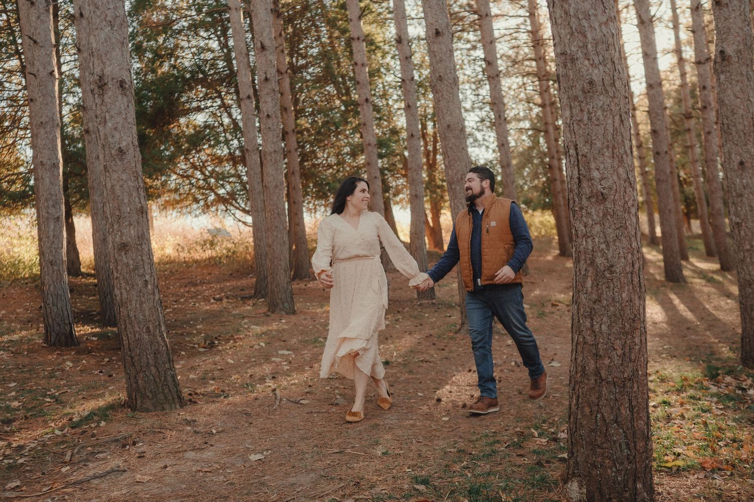 A couple holding hands while walking through a sunlit pine forest on an autumn afternoon.