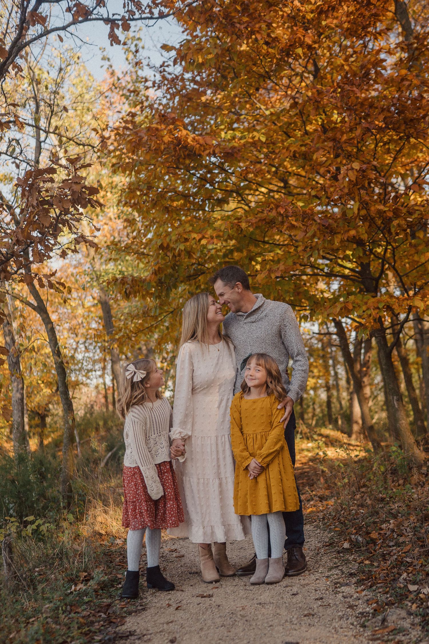 A family in coordinating autumn outfits stands together among trees with golden fall foliage.