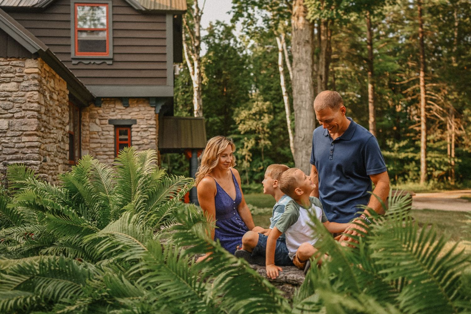Family relaxing together among ferns outside their stone and wood cabin.