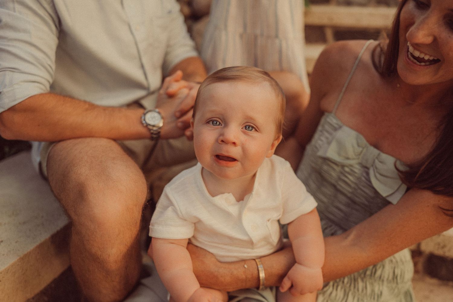 Close-up candid shot of a baby smiling while sitting on stone steps with parents.