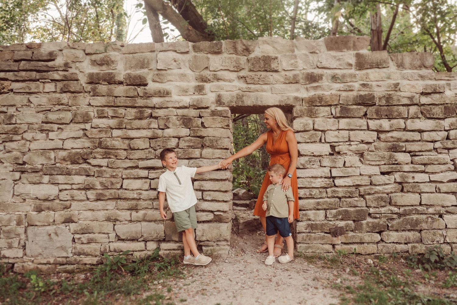 People explore an old stone wall pathway surrounded by trees and dappled sunlight.