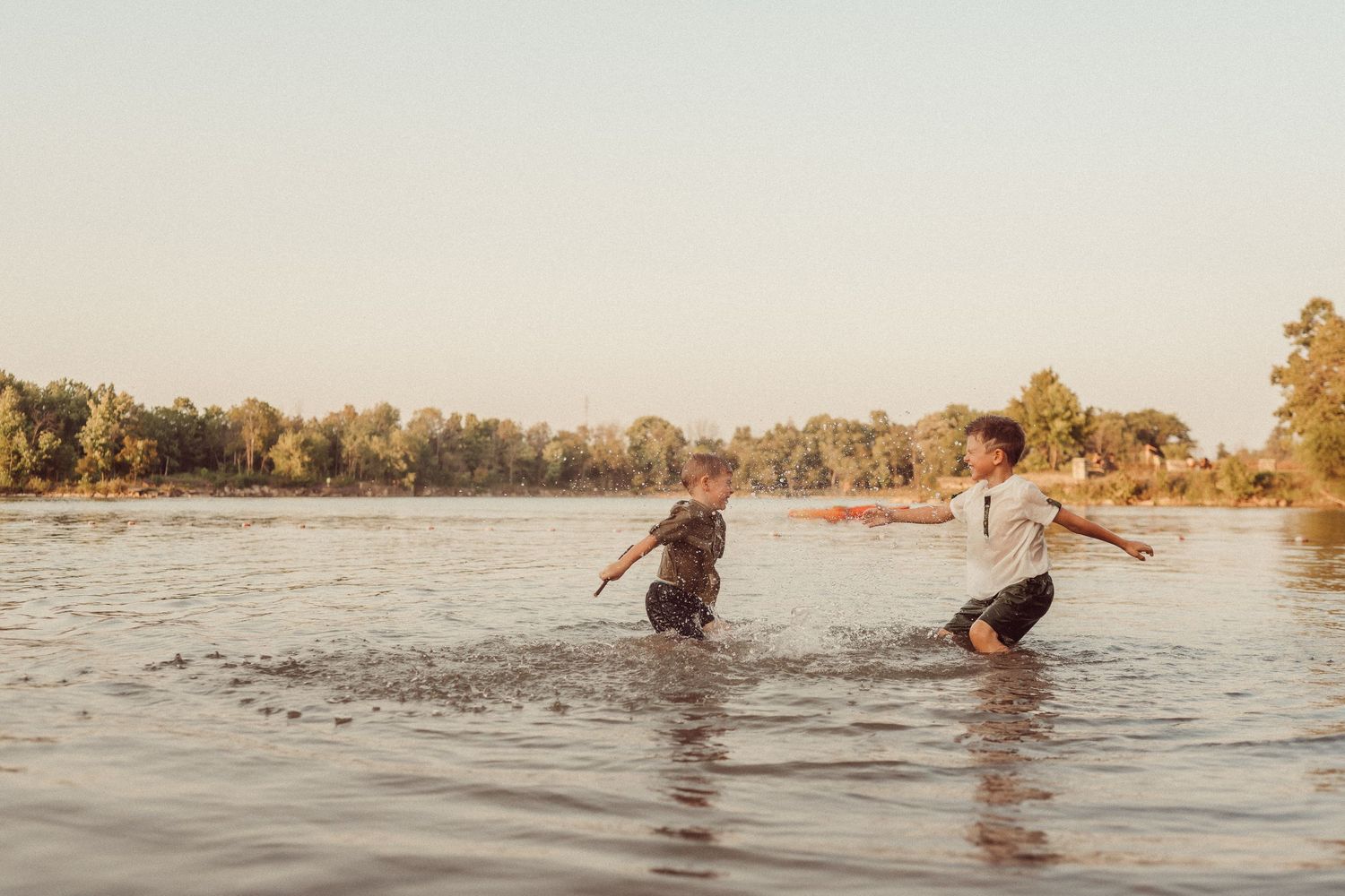 People playing and splashing in a lake during sunset with trees lining the shore.