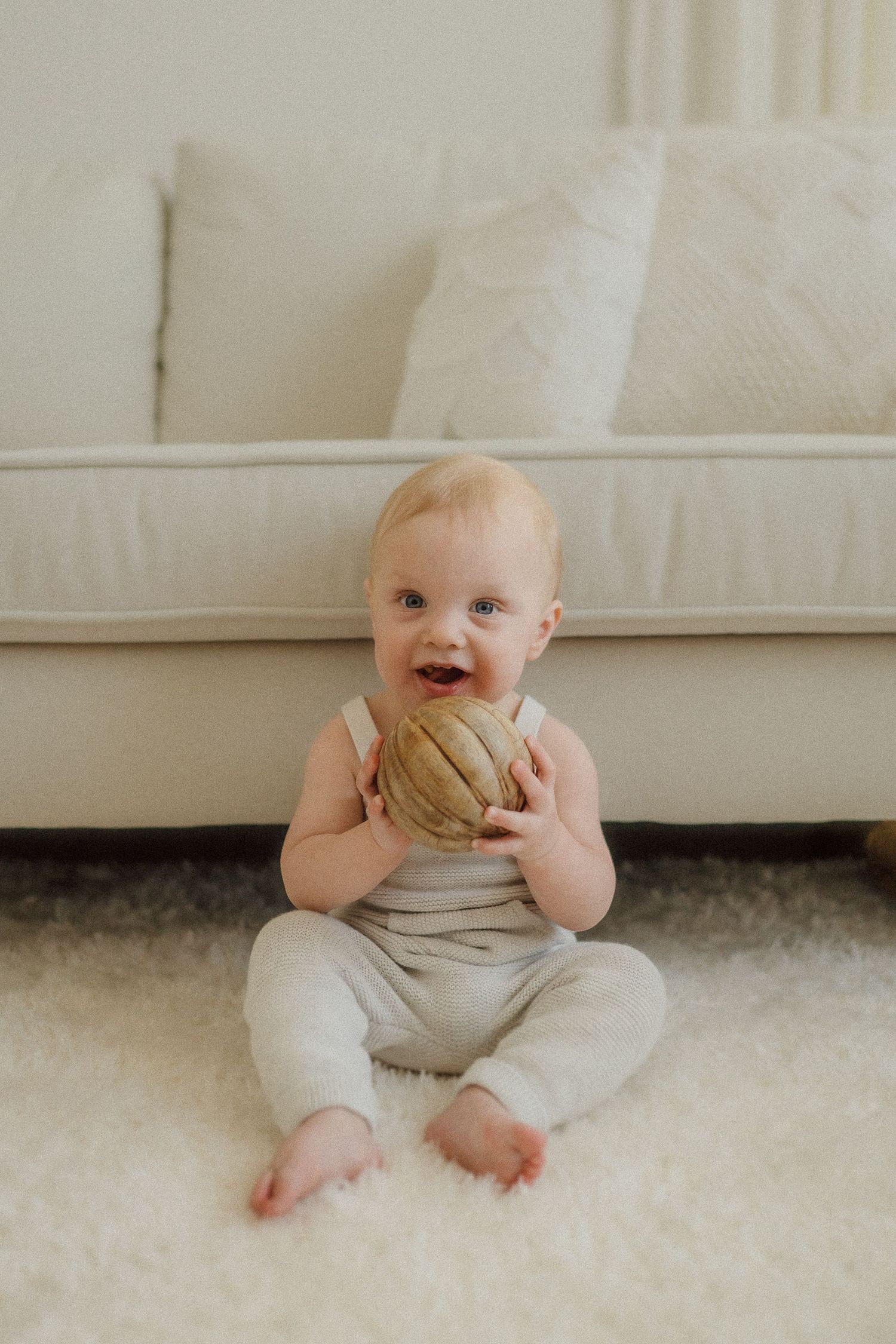 A happy baby wearing a light-colored romper sits on a plush white carpet holding a round ball.