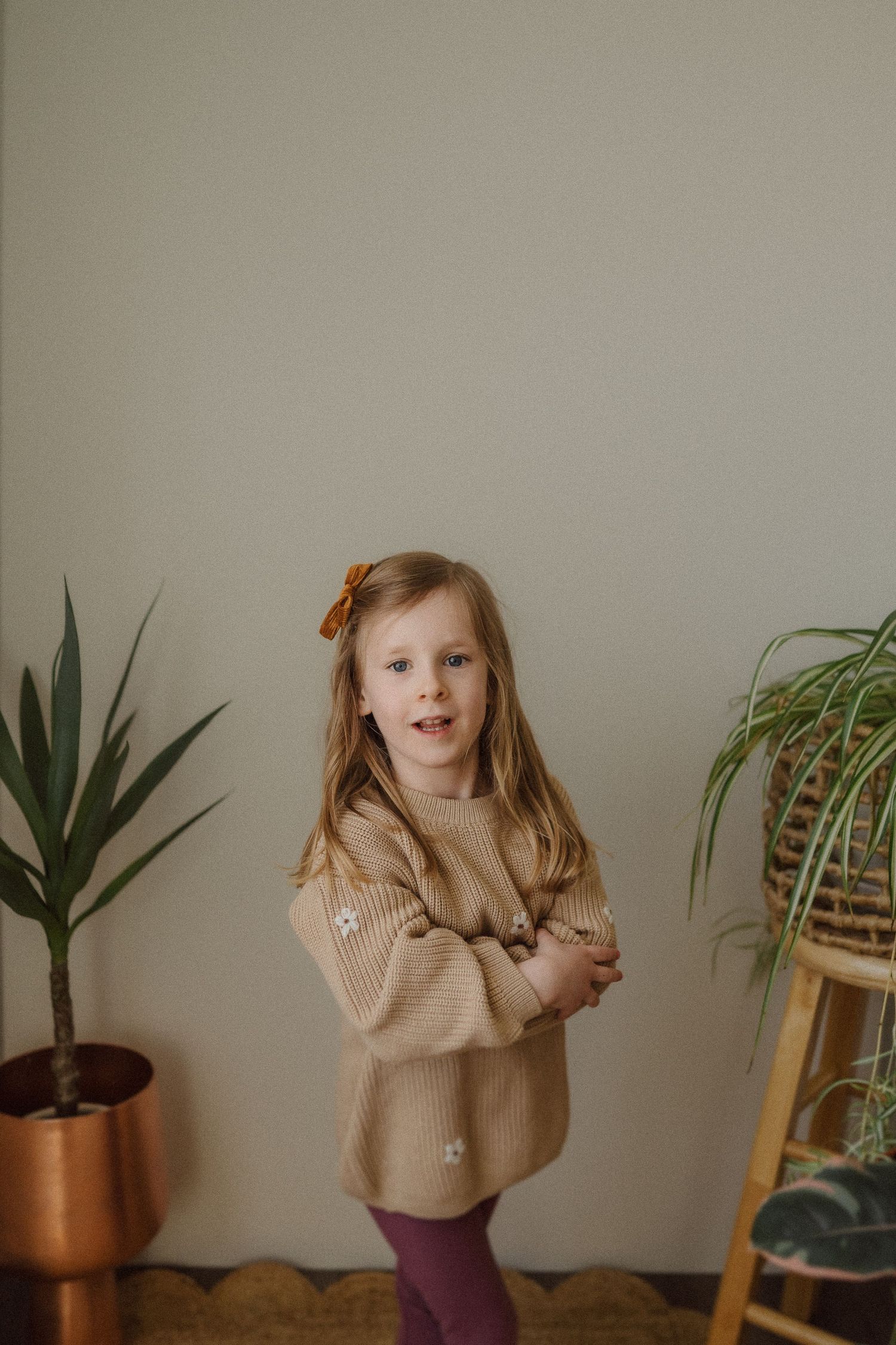 Child in oversized beige sweater poses against wall with indoor plants in modern minimal setting.