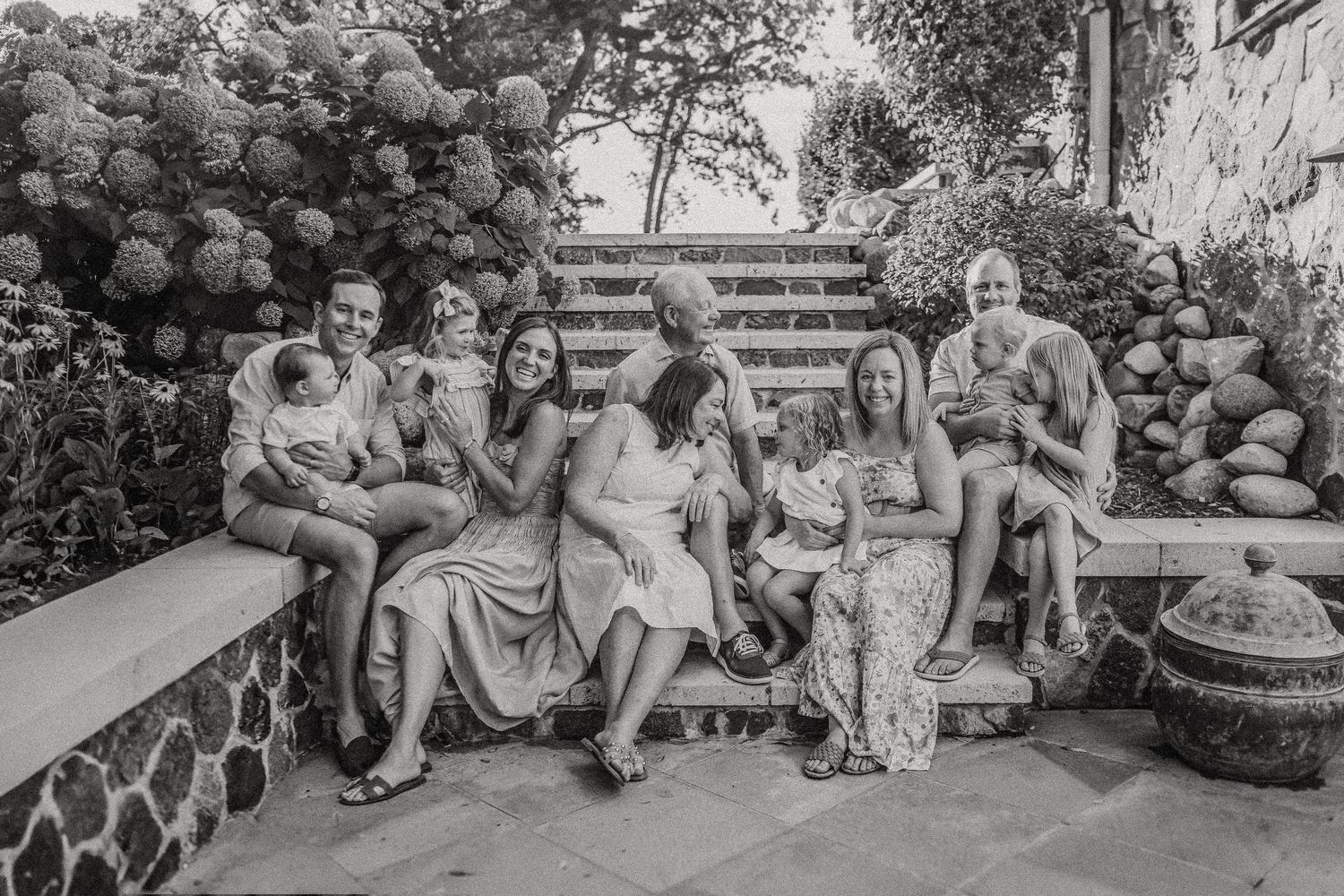 Black and white photo of family members relaxing on outdoor stone steps with flowering bushes.