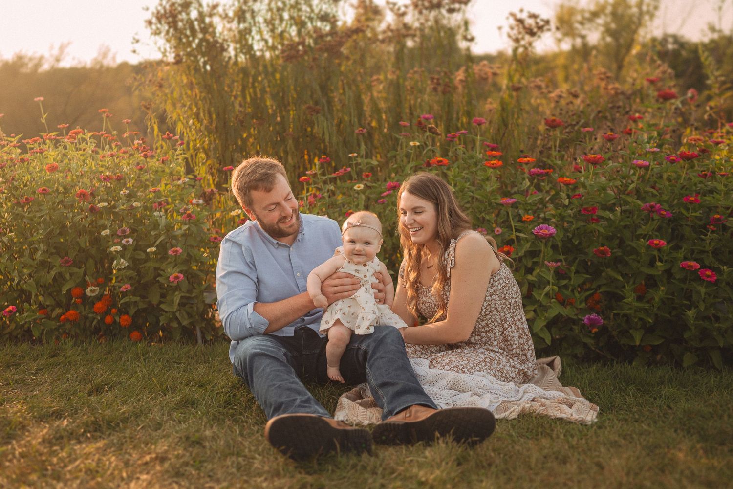 Family shares tender moment together on picnic blanket surrounded by red flowers at sunset.