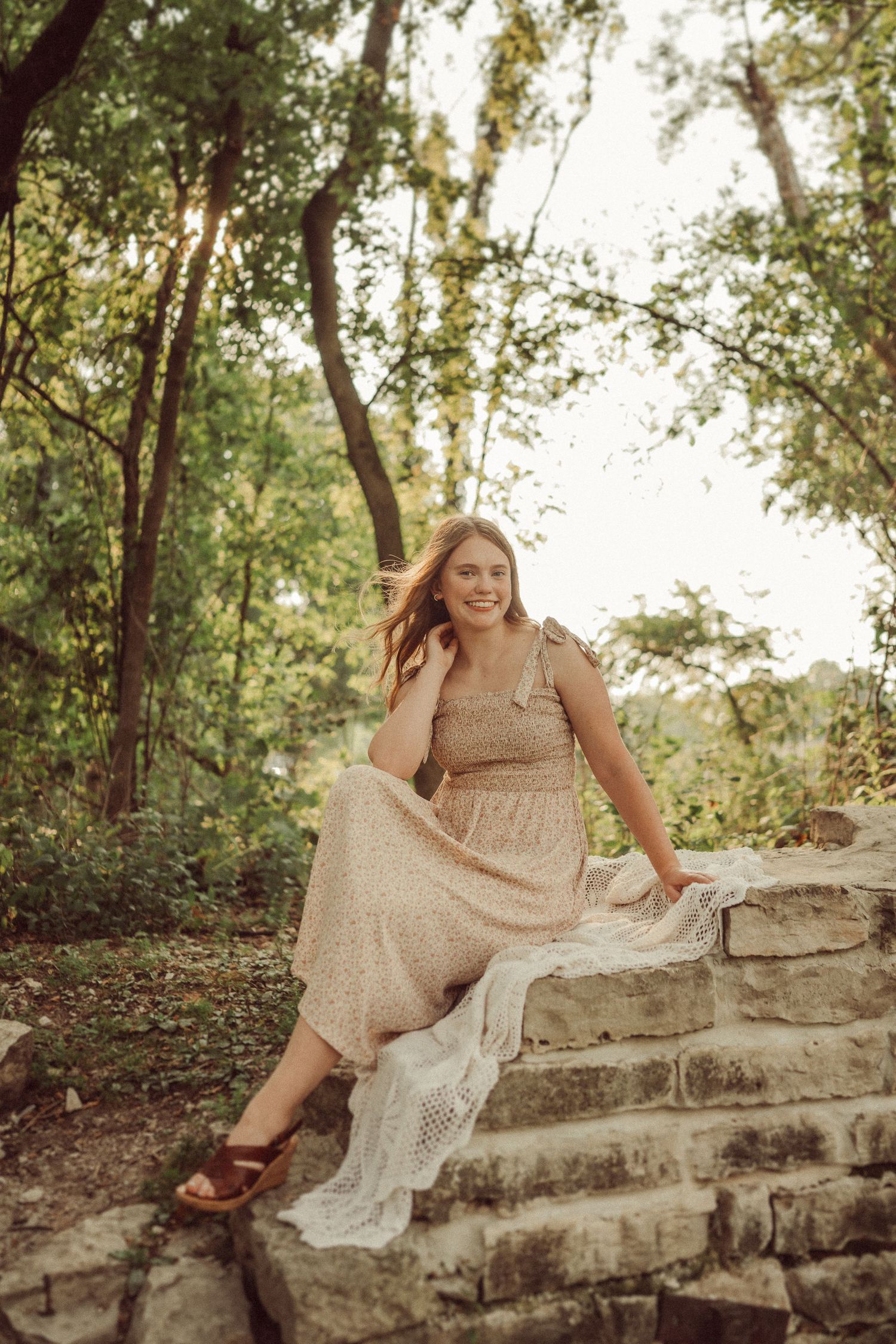 Woman in flowy dress sits casually on stone steps surrounded by lush greenery.