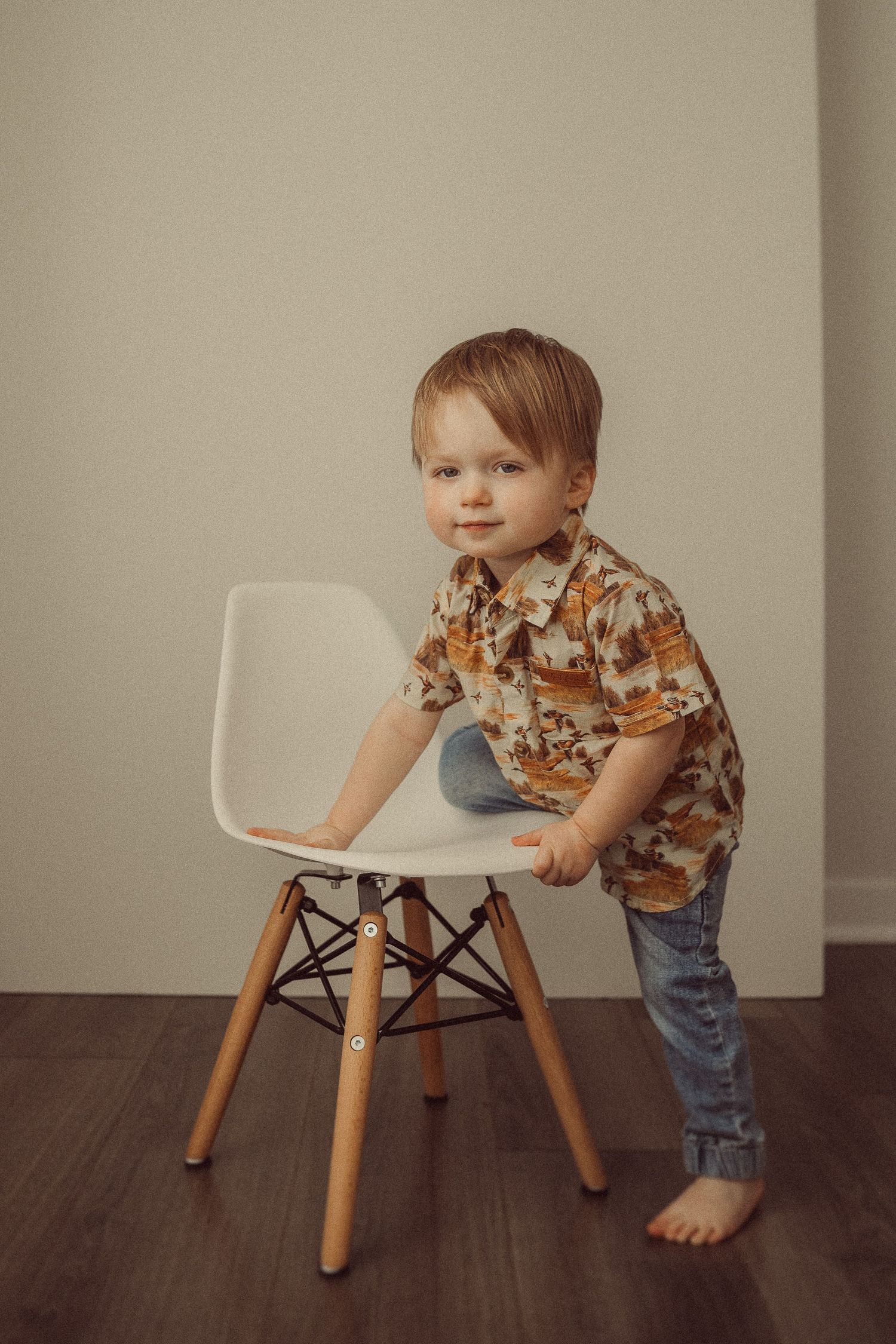 Barefoot toddler in floral shirt and jeans leaning on modern white chair.