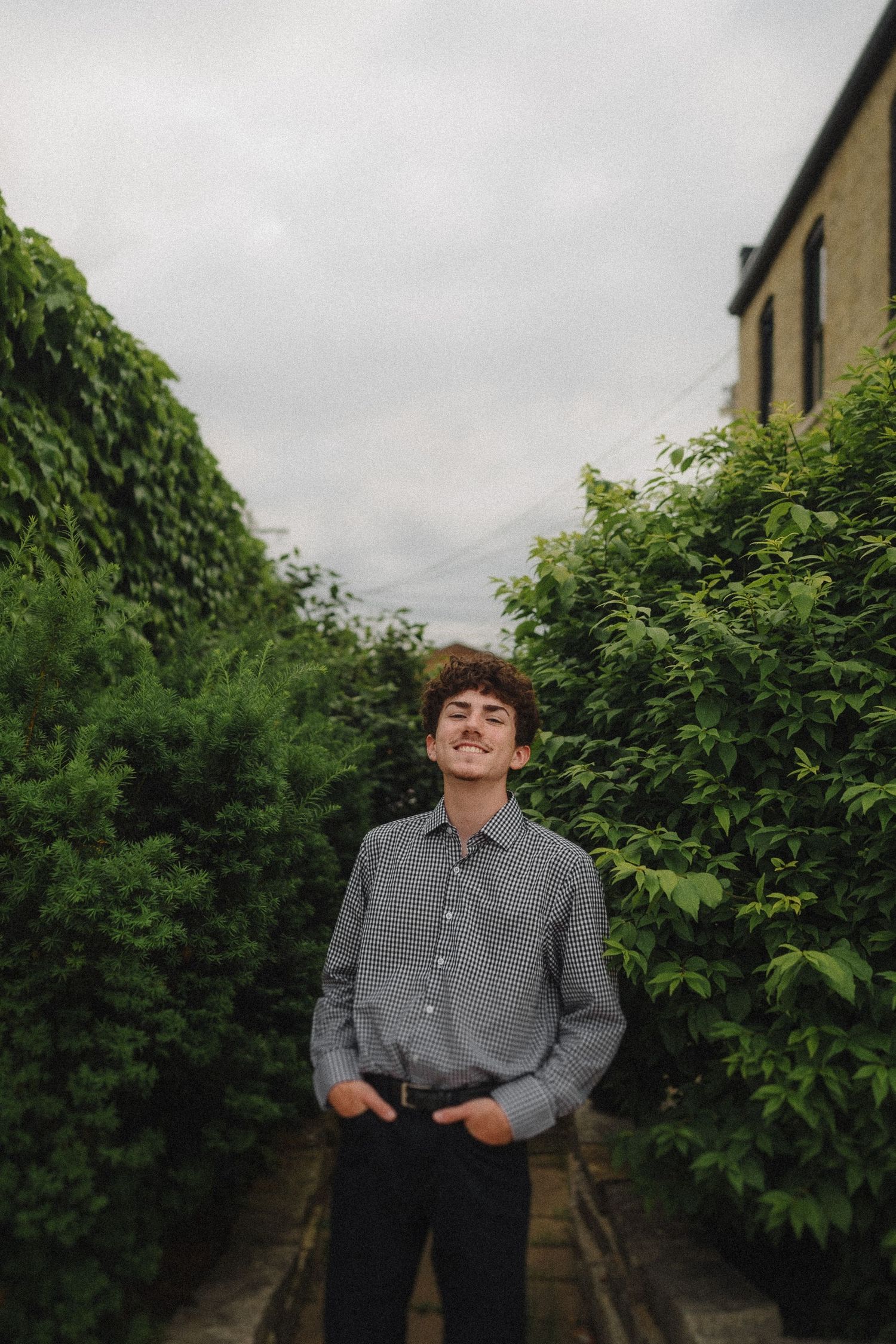 A person stands in a garden path wearing a checkered button-down shirt and dark pants against lush green foliage.