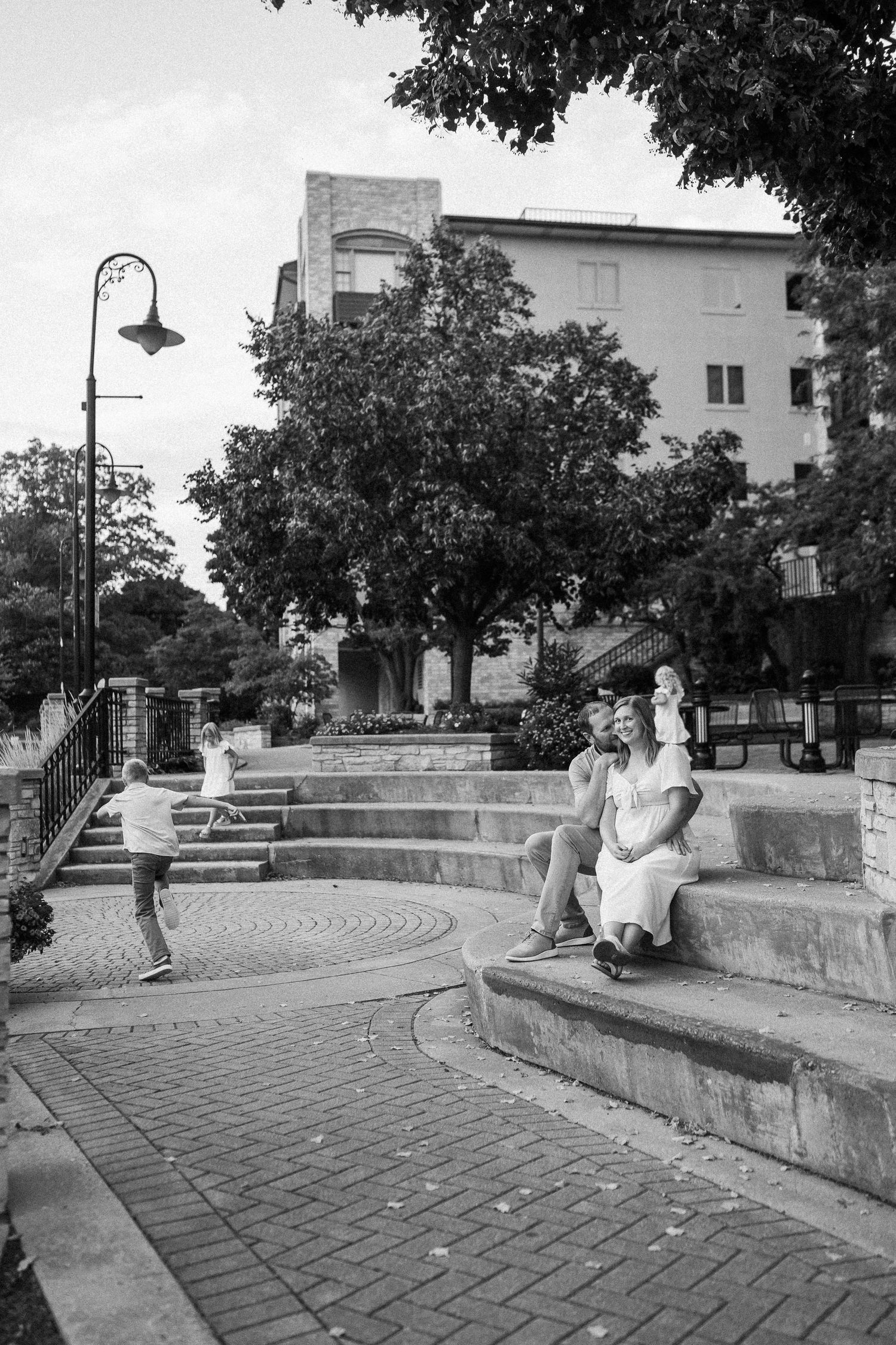 A curved brick path leads through an urban park area with benches and trees providing shade.