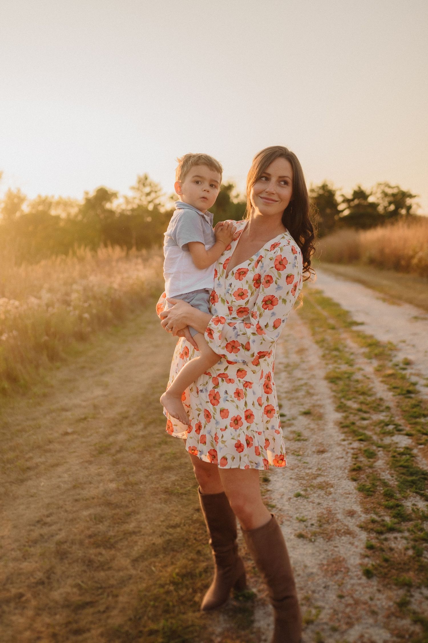 A heartwarming mother-child portrait in a floral dress against a sunset-lit rural backdrop.