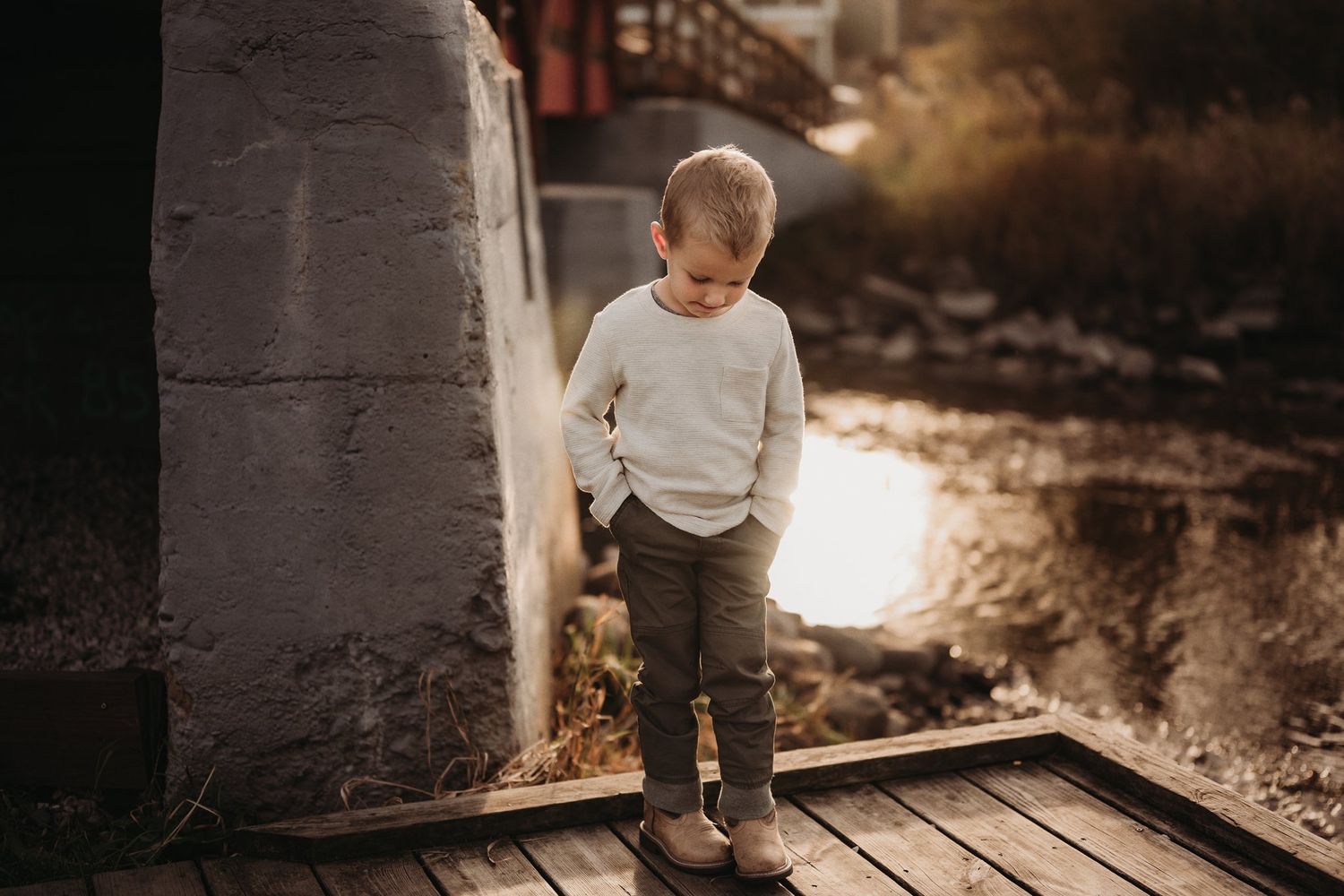 A child in a cream sweater standing on wooden planks during golden hour near water.