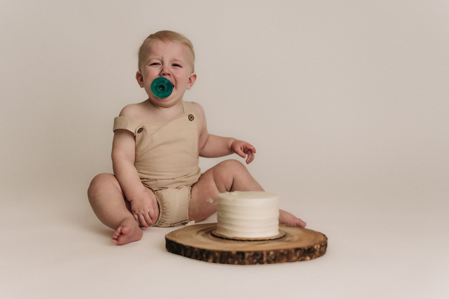 Baby in beige romper with pacifier sits next to white cake on wooden slice against neutral backdrop.