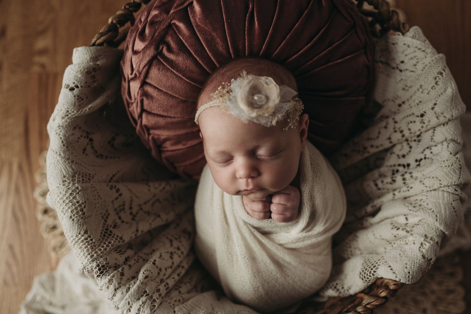 Sleeping newborn in cream wrap with floral headband resting in plush mauve cushioned bassinet.