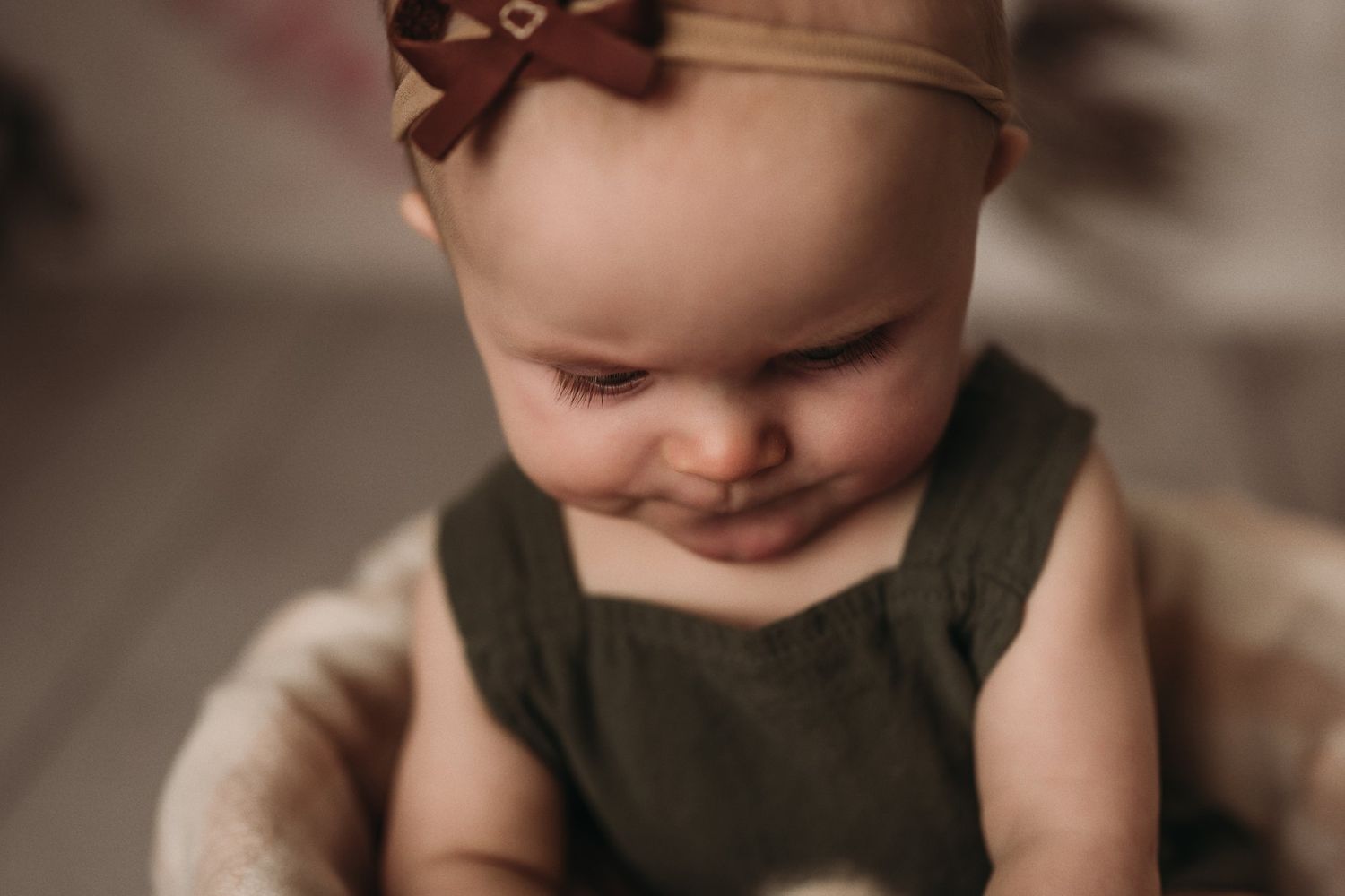 Close-up portrait of a young child wearing an olive green romper and brown bow headband against a blurred background.