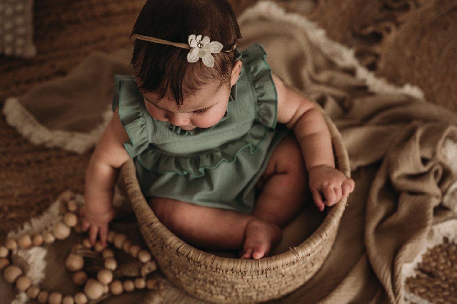 A baby wearing a sage green ruffle dress sits in a woven basket surrounded by neutral decor.