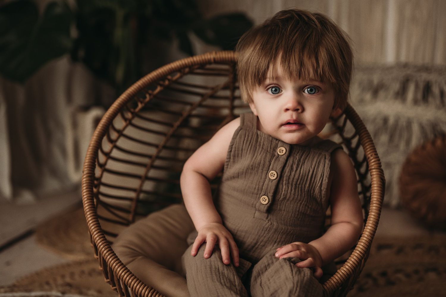 A toddler in brown clothing sits in a round wicker chair against a macrame backdrop.