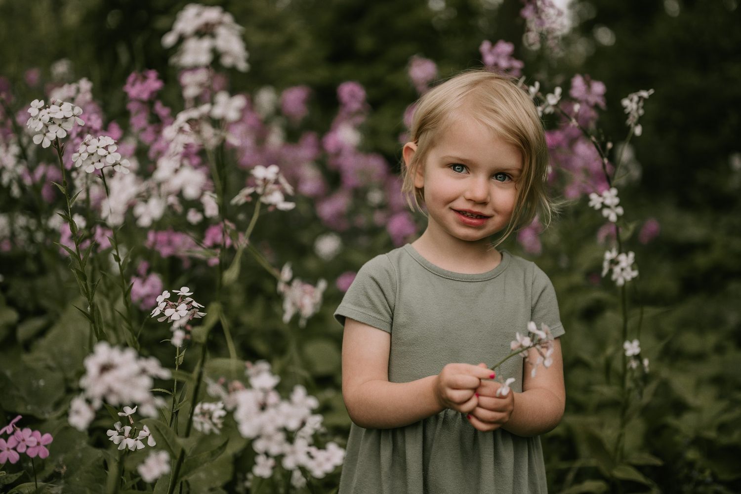 Young child in gray dress stands among pink and white wildflowers in garden setting.