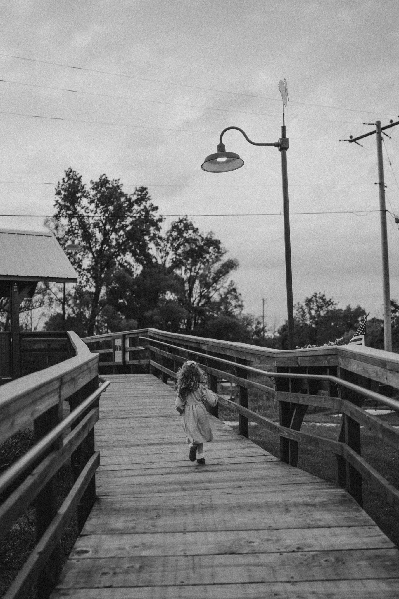 A black and white photo of a wooden boardwalk with a streetlamp against an evening sky.