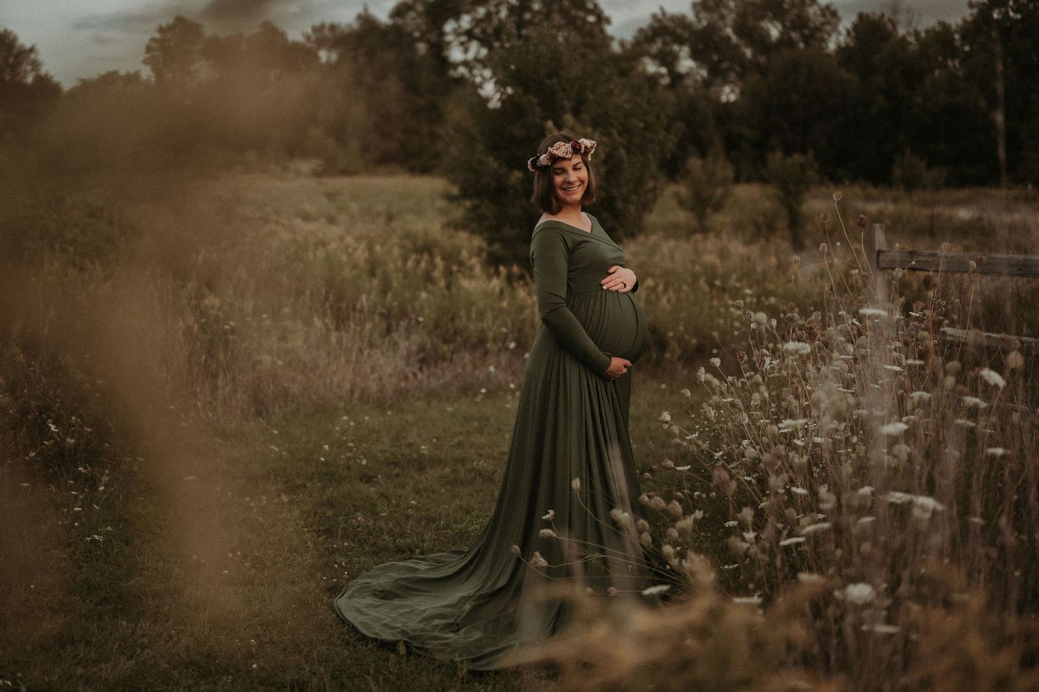 A flowing green maternity dress trails behind as the wearer stands in a rustic field.