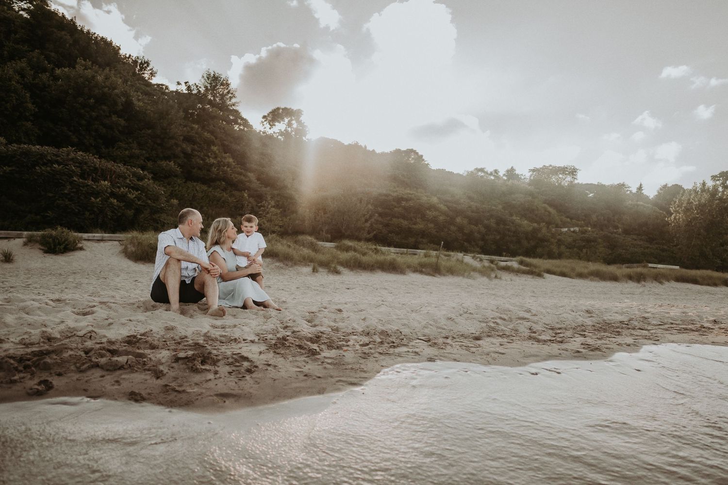 A golden sunset illuminates figures relaxing on a sandy beach with hills in the background.
