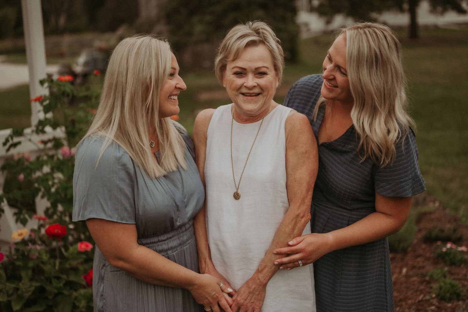 Three women share a joyful moment together in a garden with red roses blooming in the background.