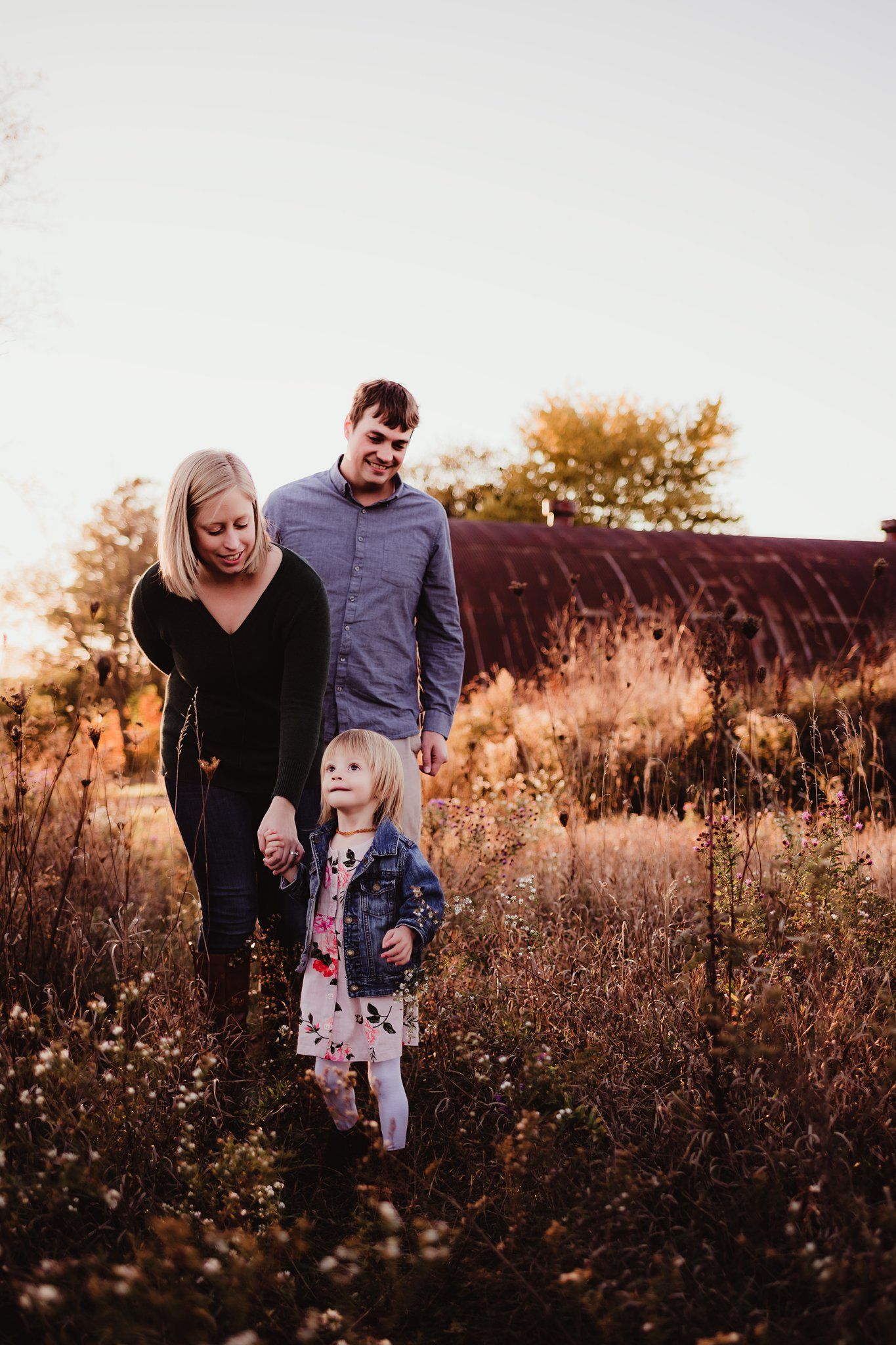 A family stands together in a golden autumn field at sunset with a rustic barn in the background.