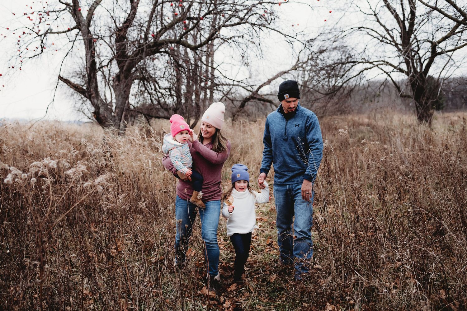 Family walking together through tall grass and bare trees in autumn landscape.