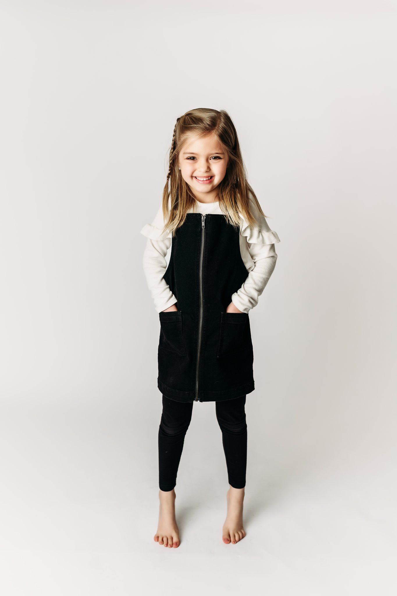 A barefoot child poses in a black zip-up dress over a white long sleeve shirt.