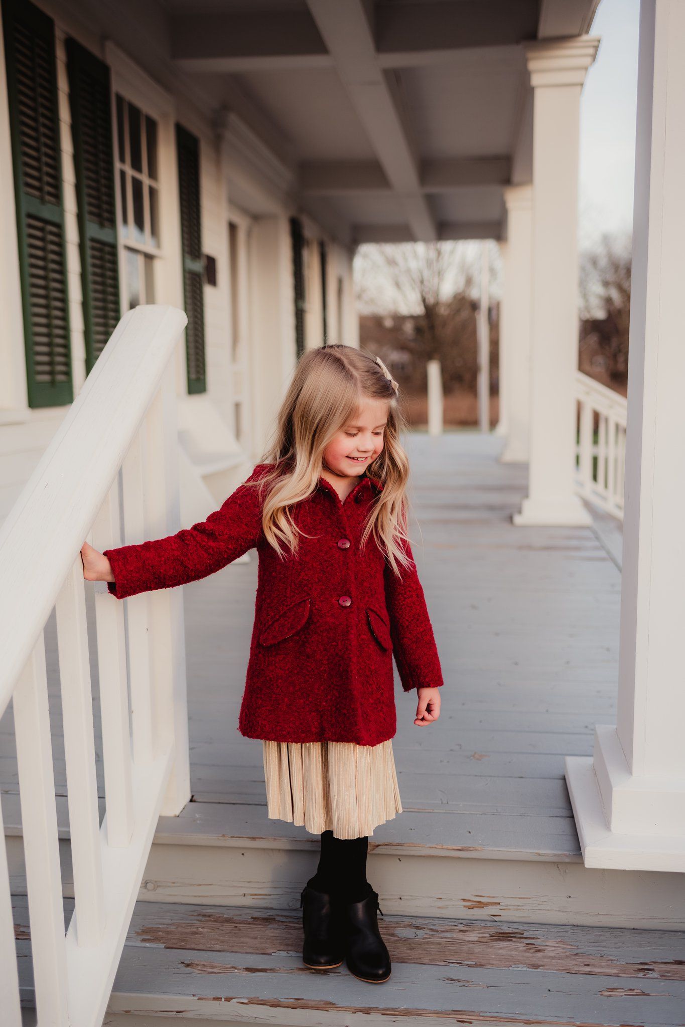 Child in red coat standing on white porch with green shutters.