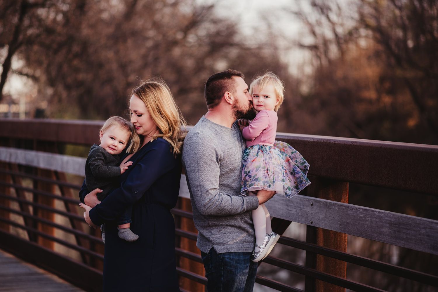 Family shares tender moment while standing on wooden bridge at sunset.