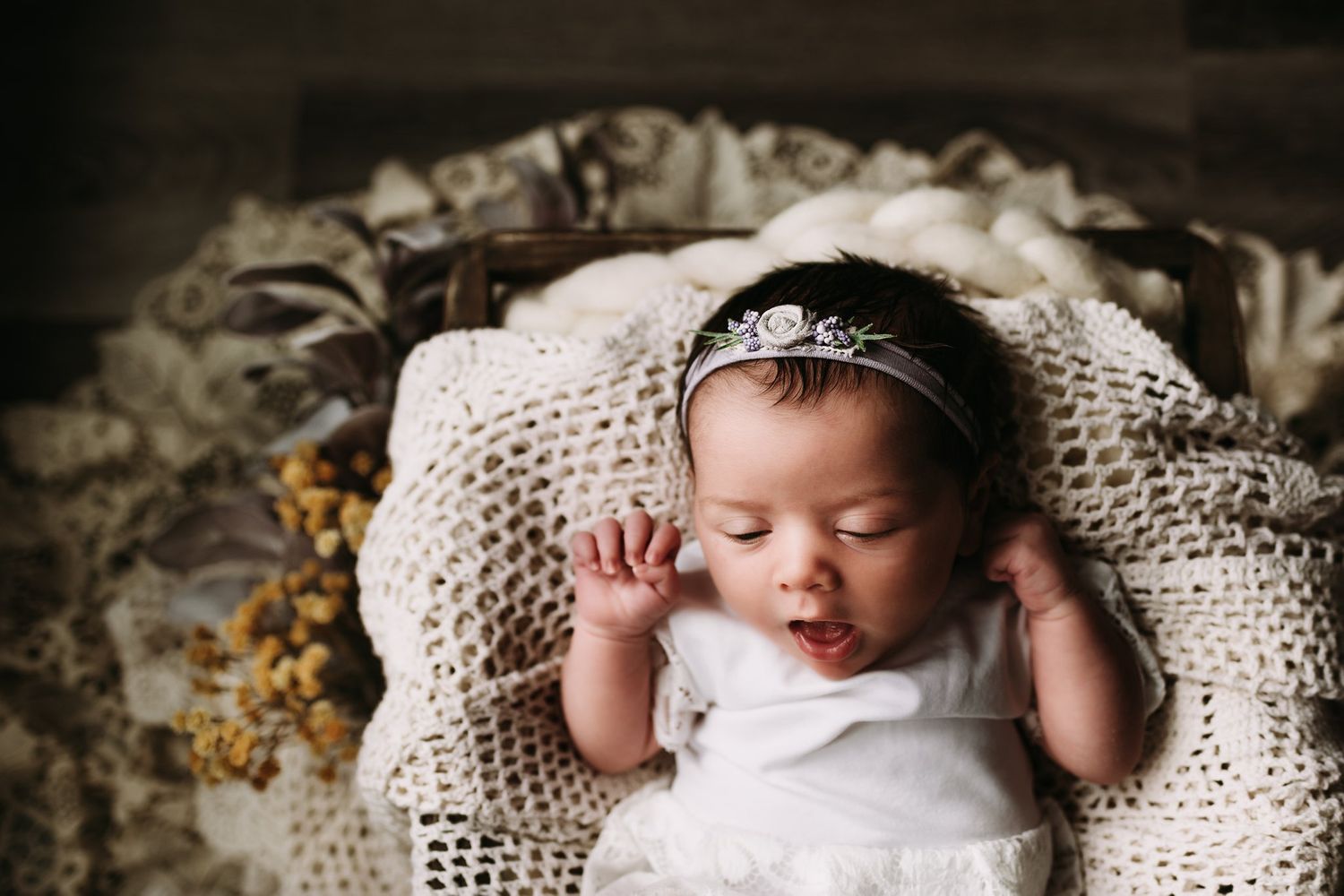 Sleeping baby in white dress and crown photographed from above on lace blanket.