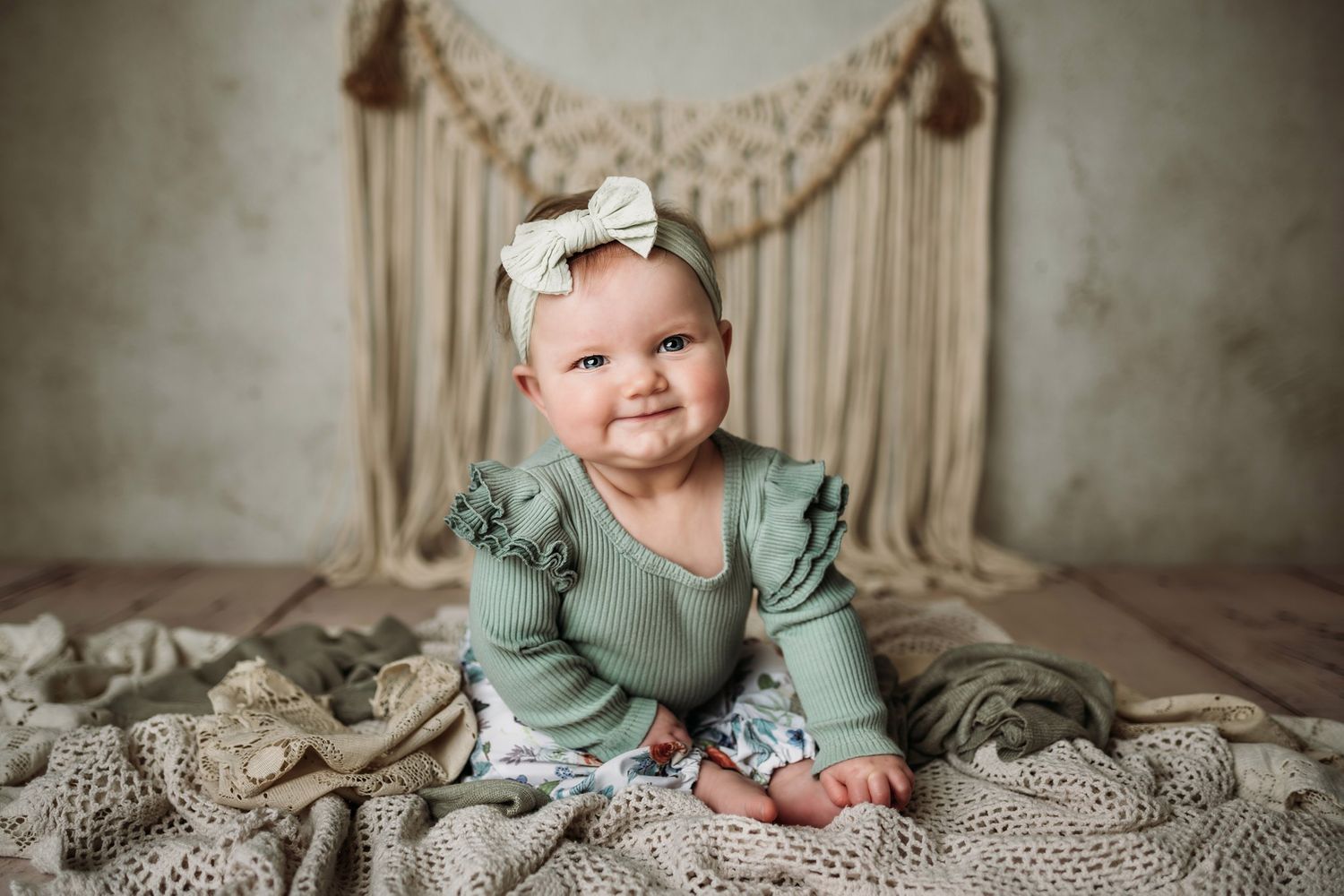Baby in mint green outfit sits on textured blanket with boho wall hanging in background.