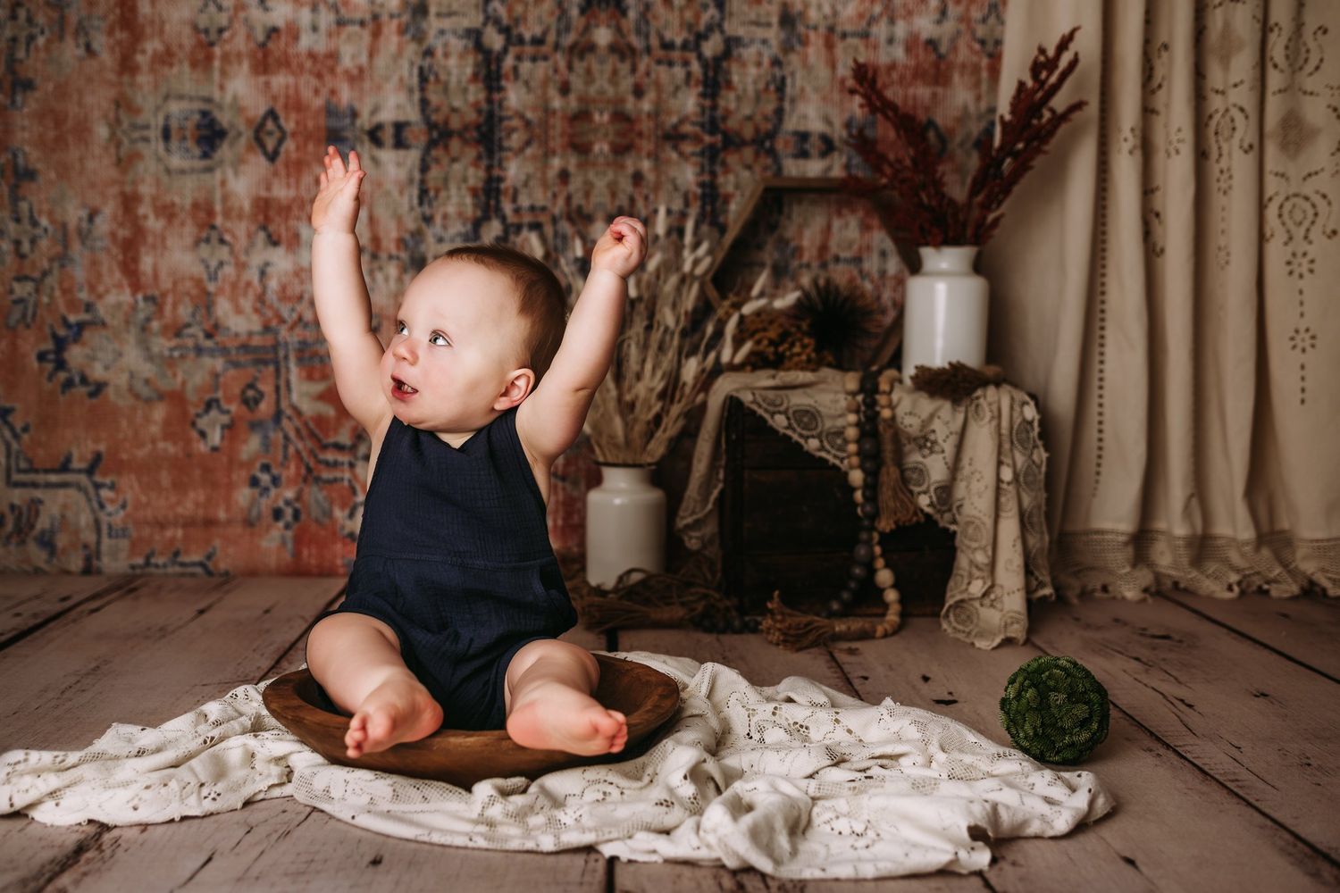 Excited baby in navy romper raises arms while sitting in wooden bowl on lace blanket.
