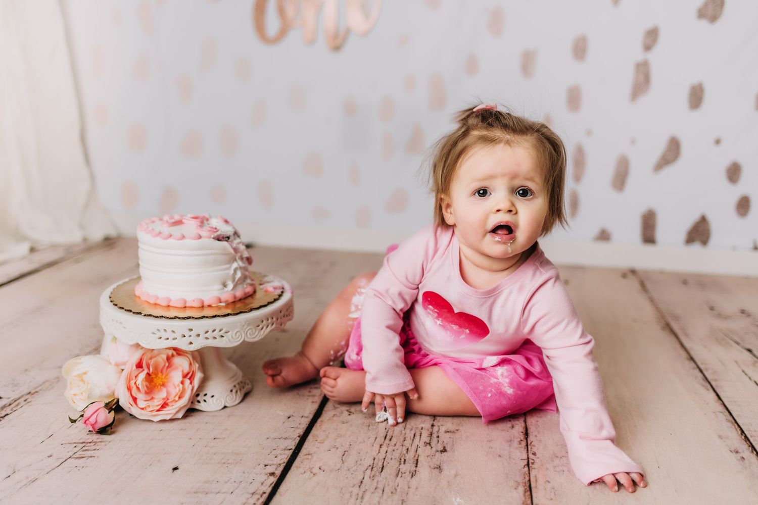 Pink-clad figure sitting by decorated birthday cake with floral accents on wooden floor.