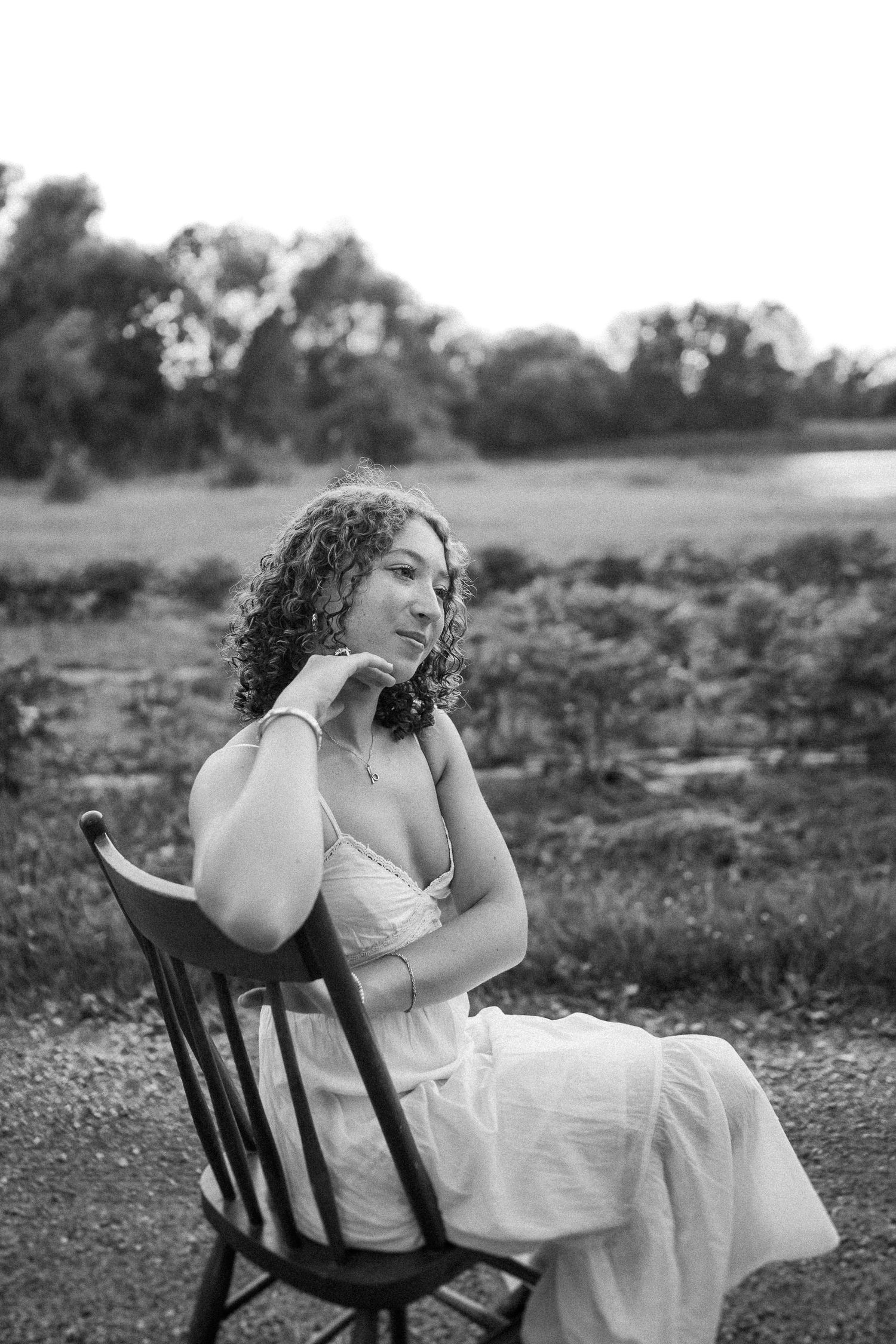 Black and white portrait of a person in a white dress seated on a wooden chair outdoors in a natural setting.