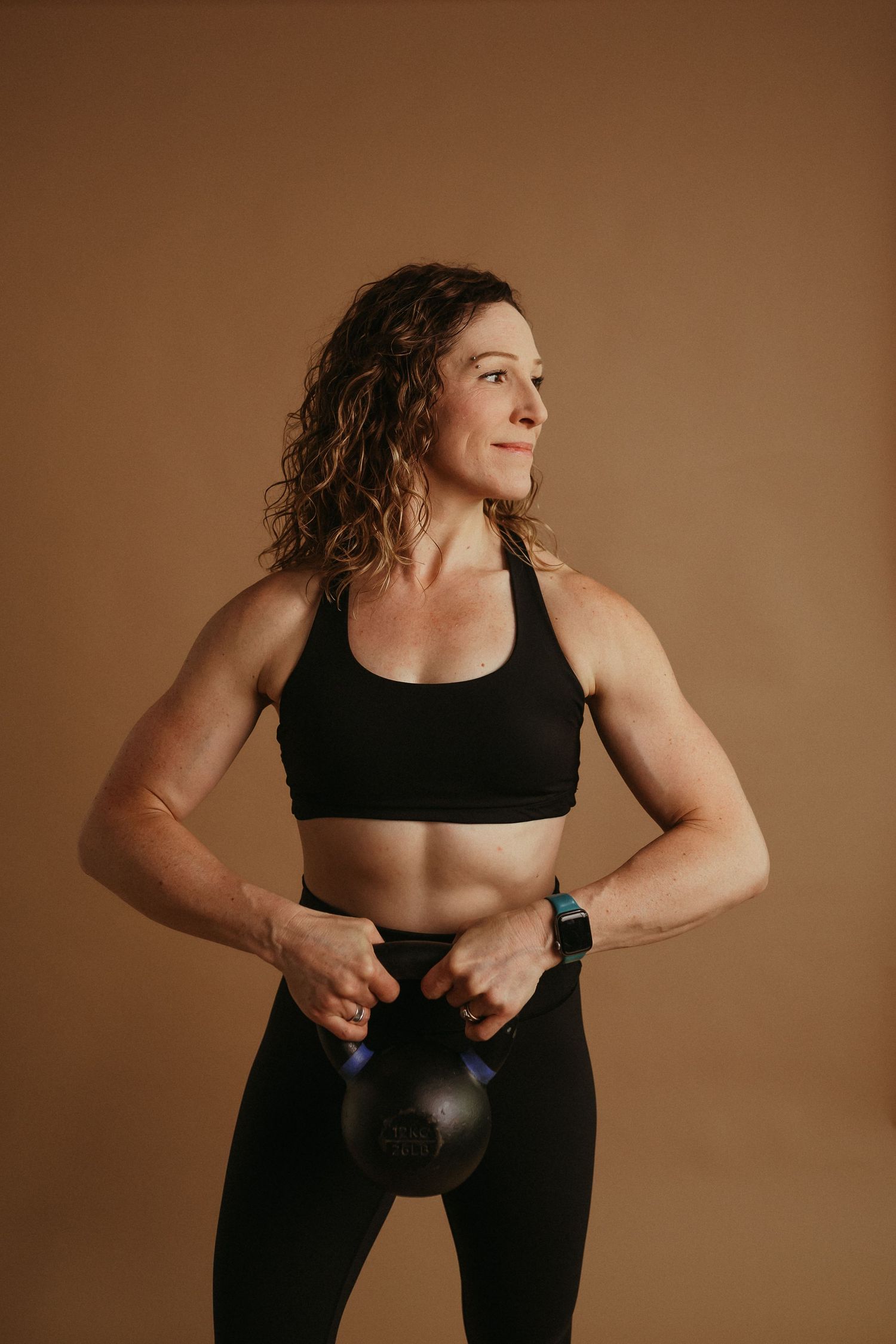 Athletic woman in black workout attire holding a kettlebell poses against brown backdrop during fitness photoshoot.