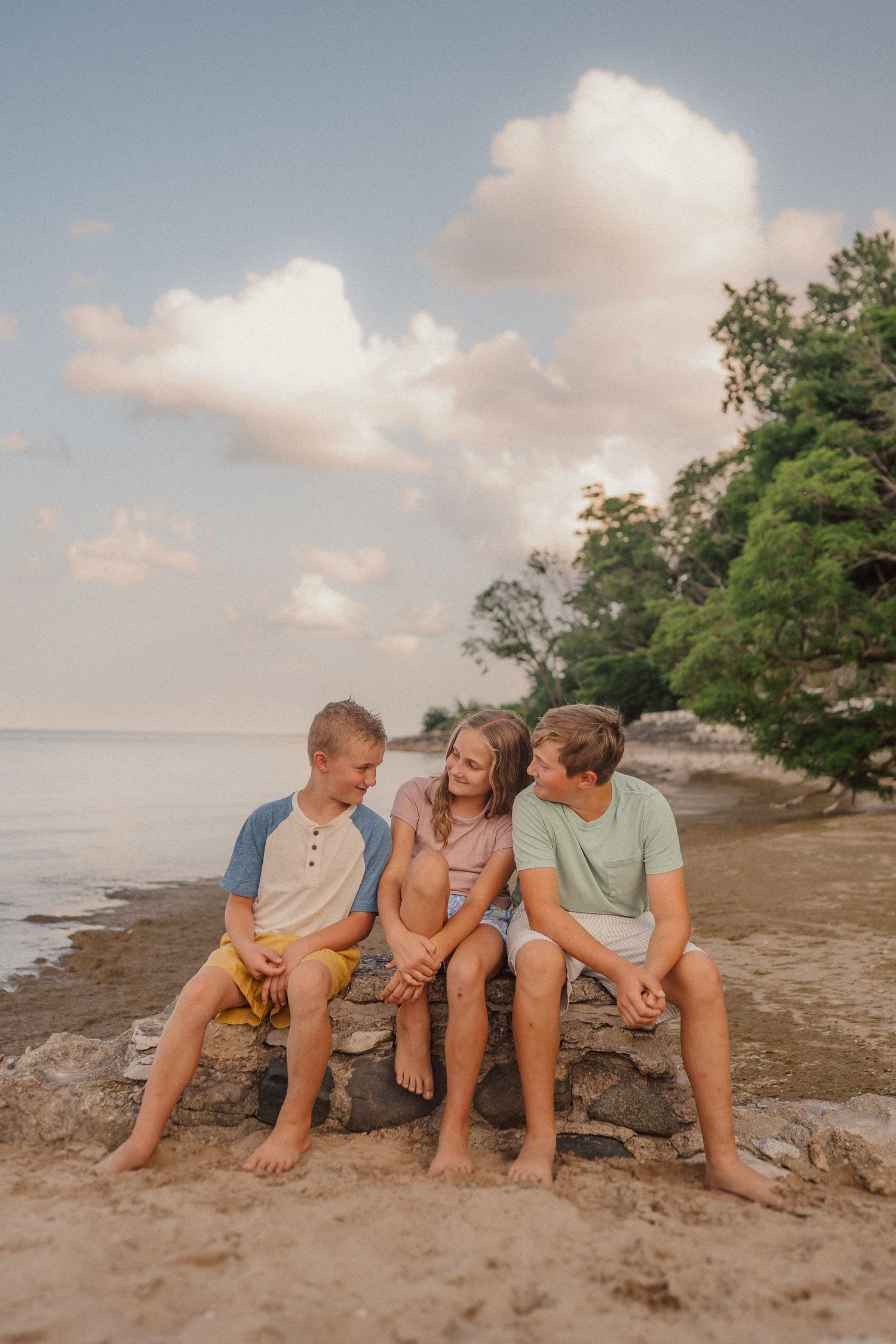 A group sits close together on a rocky beach shore with trees and cloudy skies in the background.