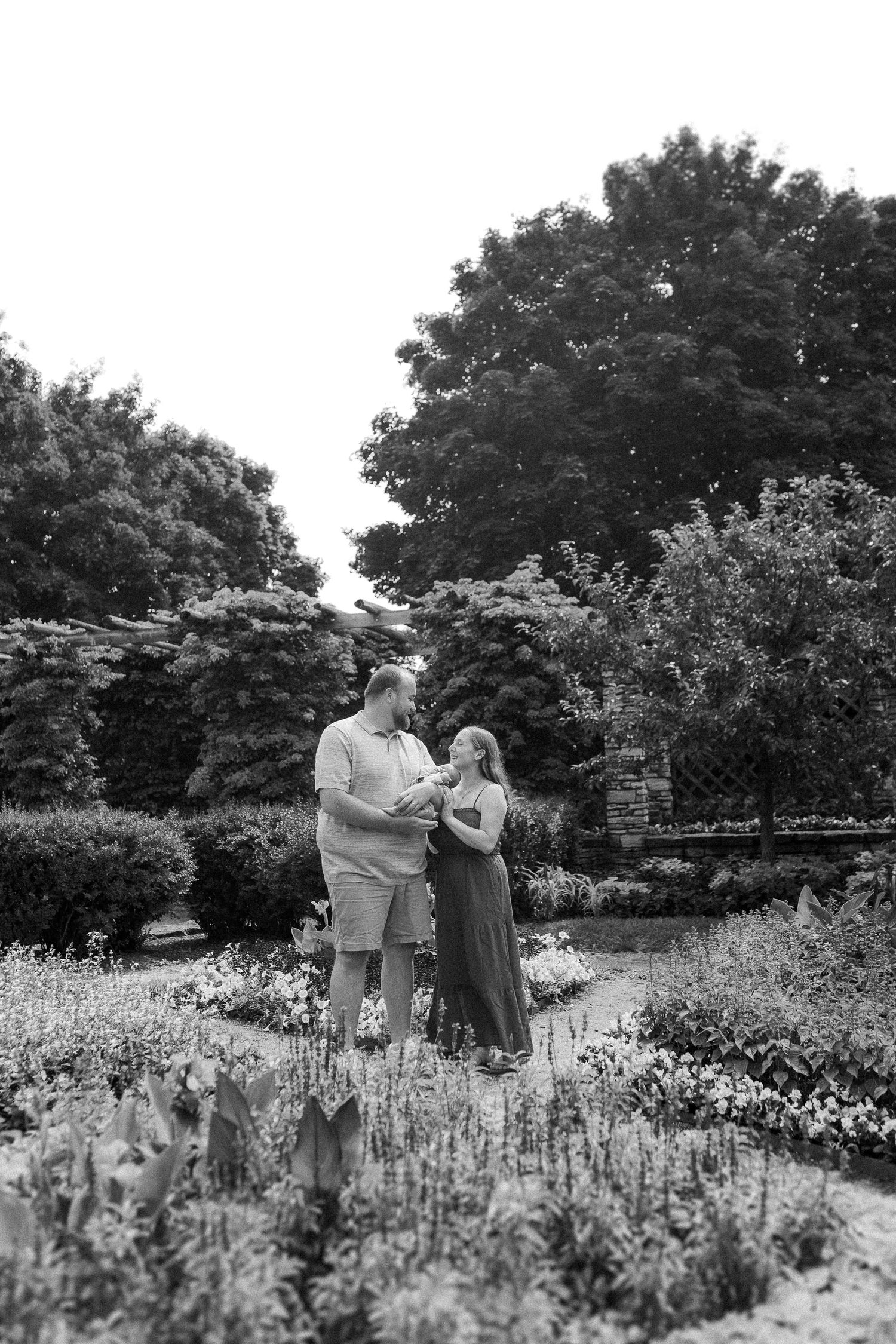 A couple shares a romantic moment while standing in a blooming garden with trees in the background, shot in black and white.
